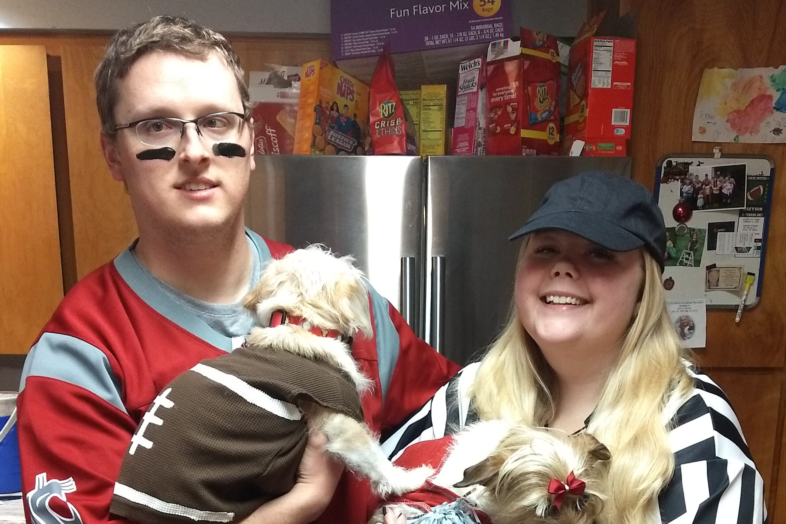 Two people pose in a kitchen with a small fluffy dog; man wears red outfit and black eye paint, woman wears a black-and-white striped top and cap, fridge and snack boxes in the background