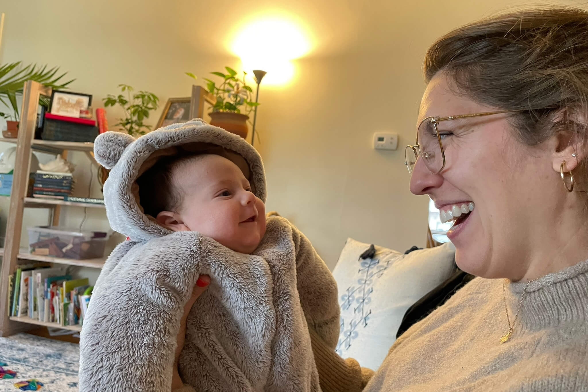 Woman wearing glasses smiles at a baby in a gray fluffy bear costume indoors, with a bookshelf and plants in the background.