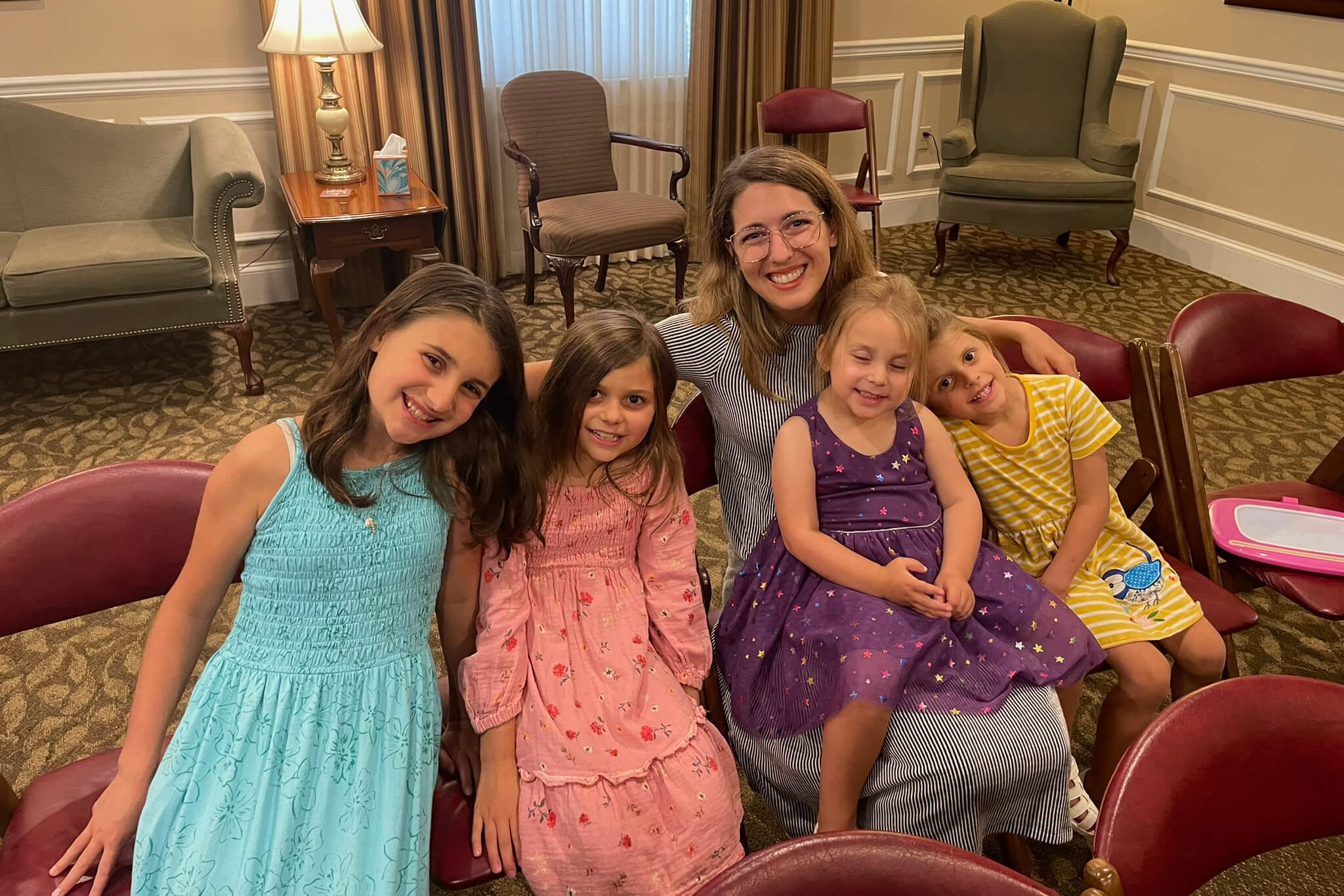 Smiling woman with four young girls in colorful dresses, posed together on red chairs in a cozy lounge.