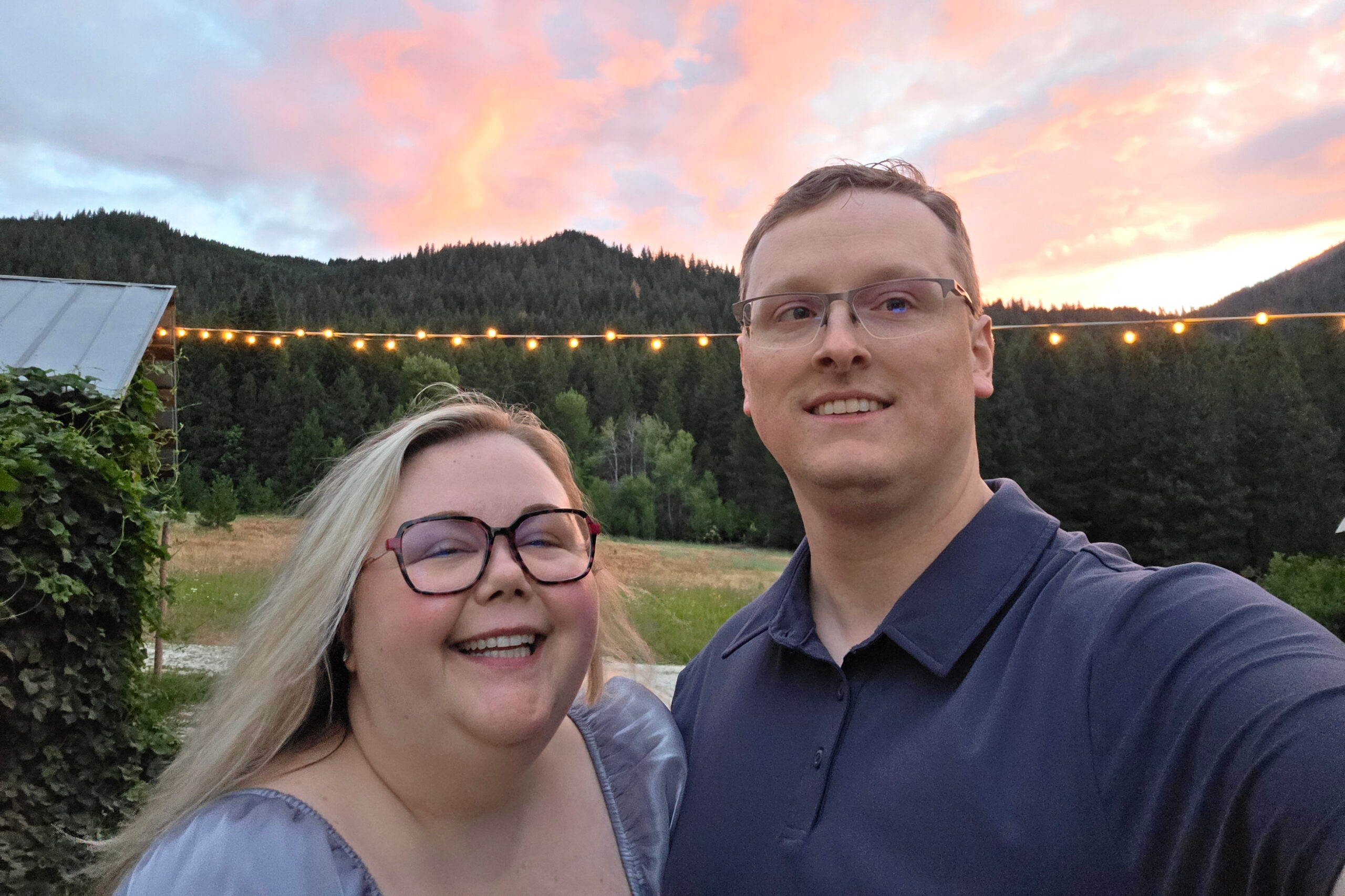 Two people smiling for a selfie outdoors at sunset with string lights and forested hills behind.