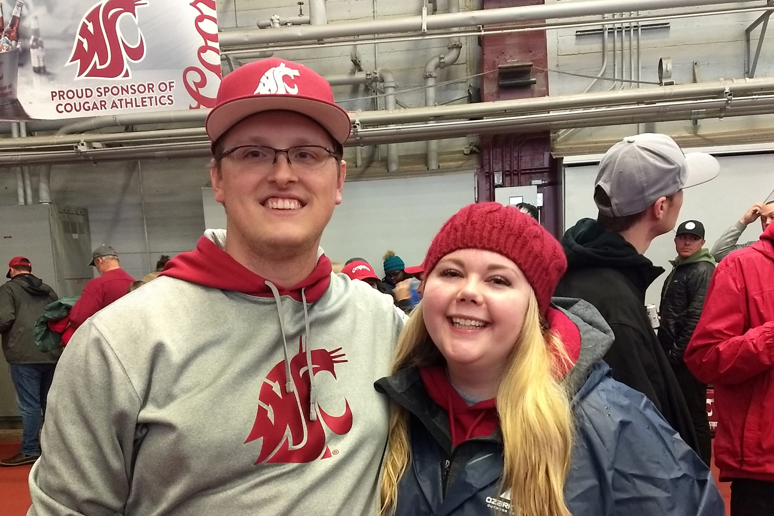 Two smiling people pose for a photo indoors at a sporting event, wearing red and gray team apparel; a banner behind them reads 'Proud Sponsor of Cougar Athletics'.