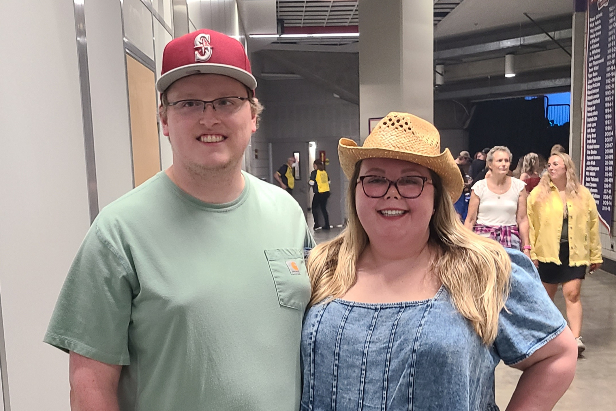 Smiling man in a green T-shirt and red cap beside a woman wearing a straw hat at an indoor event with people in the background.