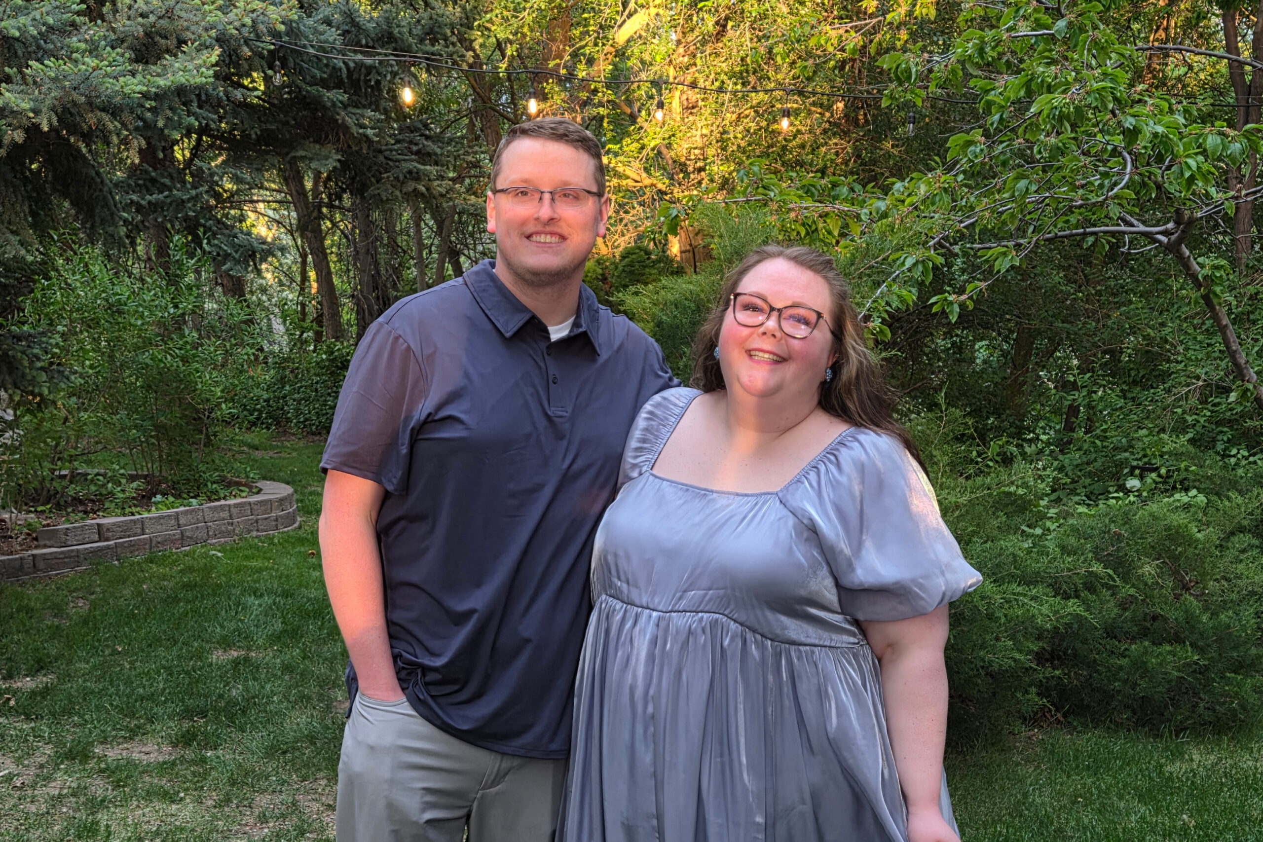 A smiling couple stands close together in a garden with green trees and string lights above them at dusk.