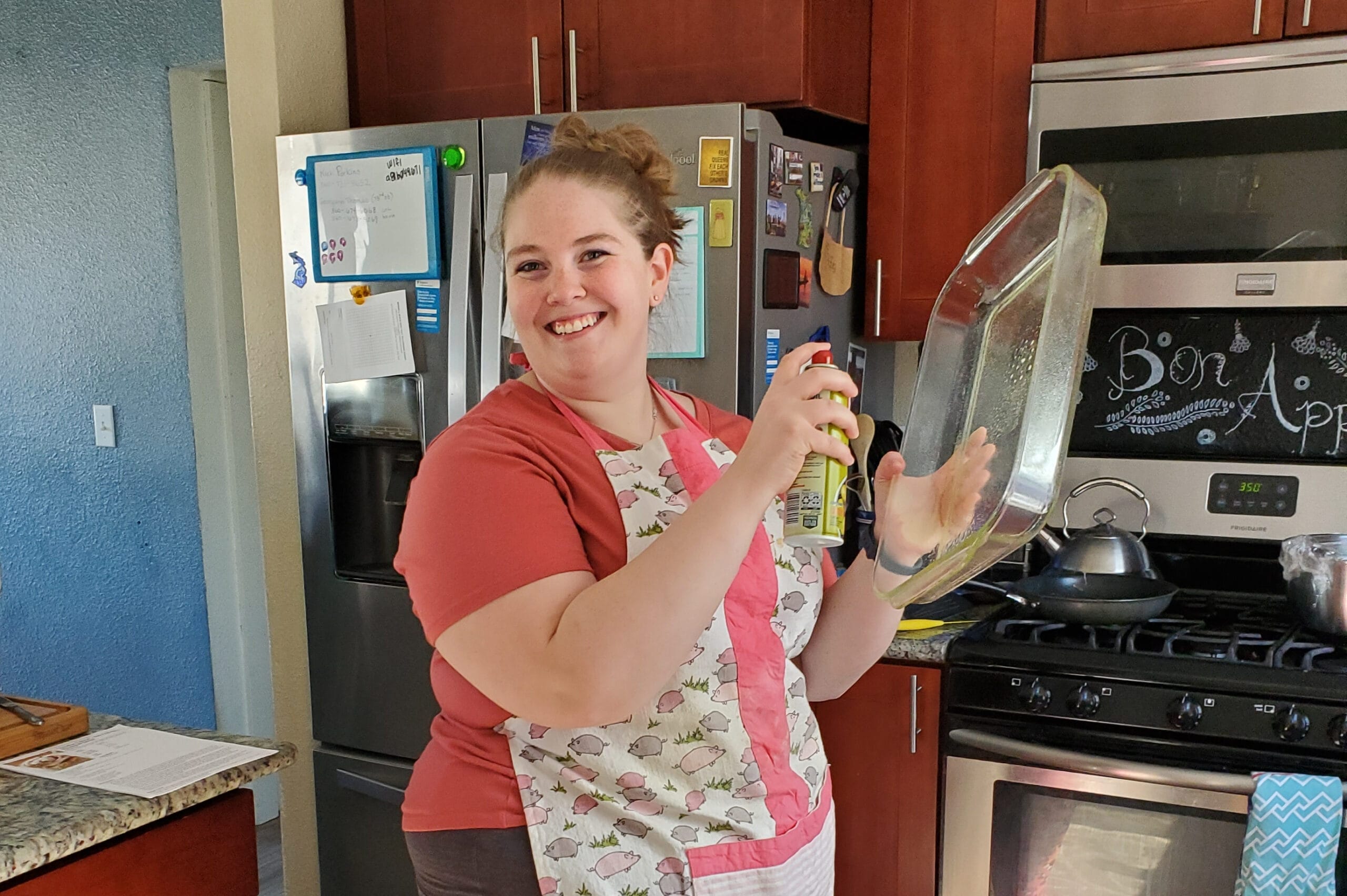 Smiling woman in a kitchen holding a spray bottle and a glass lid over a stove. She wears a coral shirt and a patterned apron.