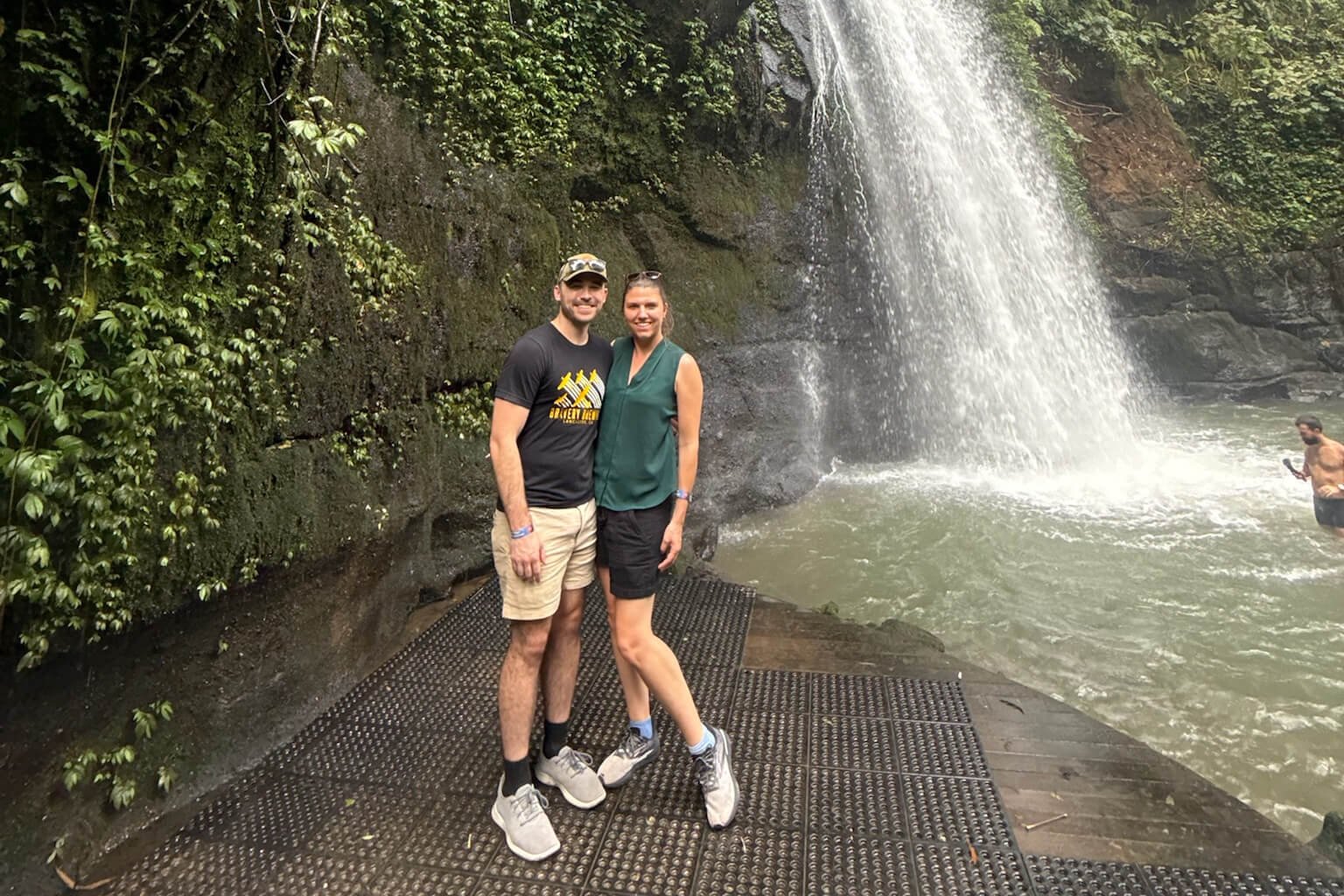 Couple posing on a metal platform by a cascading waterfall with a rocky, mossy wall and lush greenery behind them.