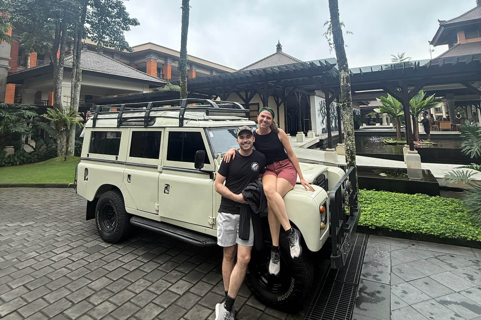 A couple smiles as they sit on the front of a white safari-style SUV parked in a resort courtyard with palm trees and water features nearby.