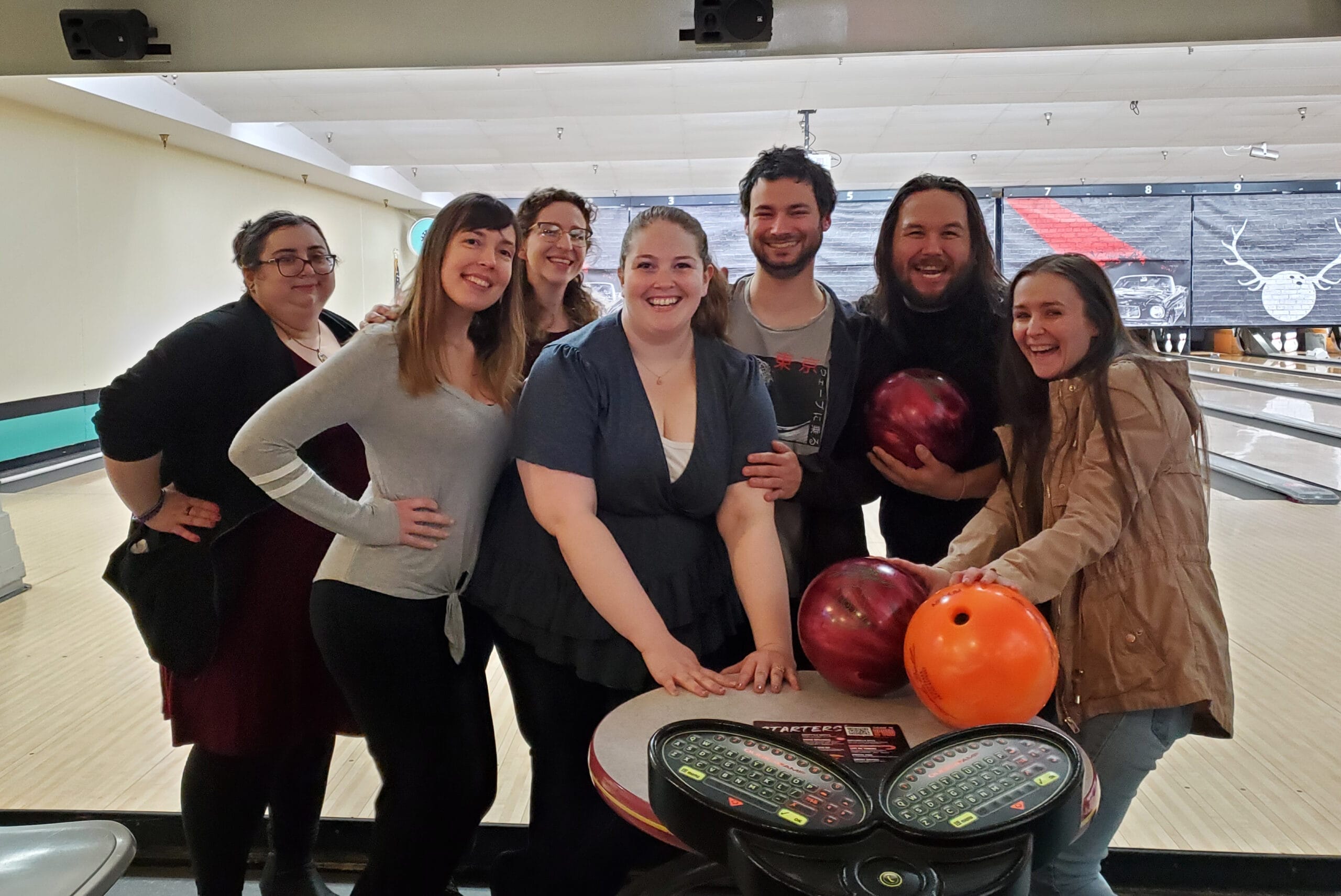 Group of seven friends smiling for a photo at a bowling alley, with bowling balls on a table in the foreground and lanes in the background
