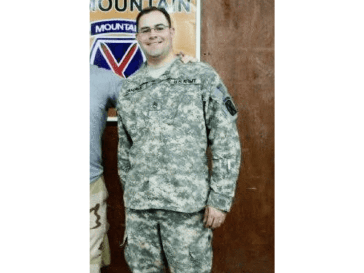 Man in Army camouflage uniform posing indoors, with a poster in the background.
