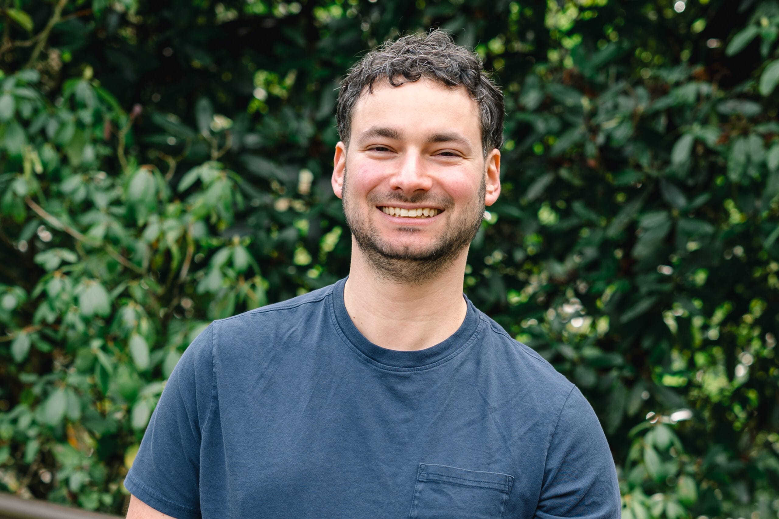Smiling man in a blue t-shirt standing outdoors with green foliage in the background, facing camera.