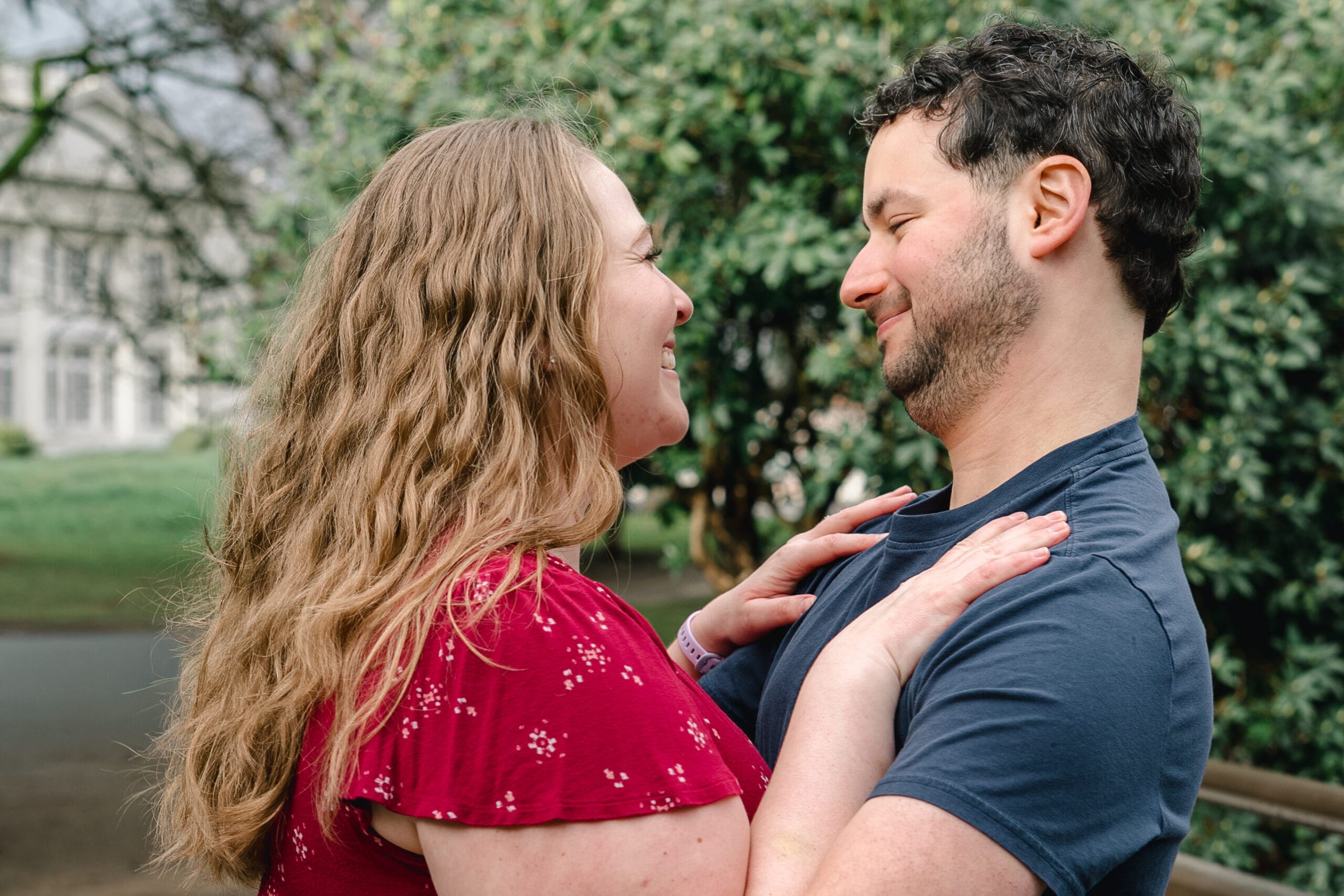 Couple hugging and smiling at each other outdoors, with greenery in the background.