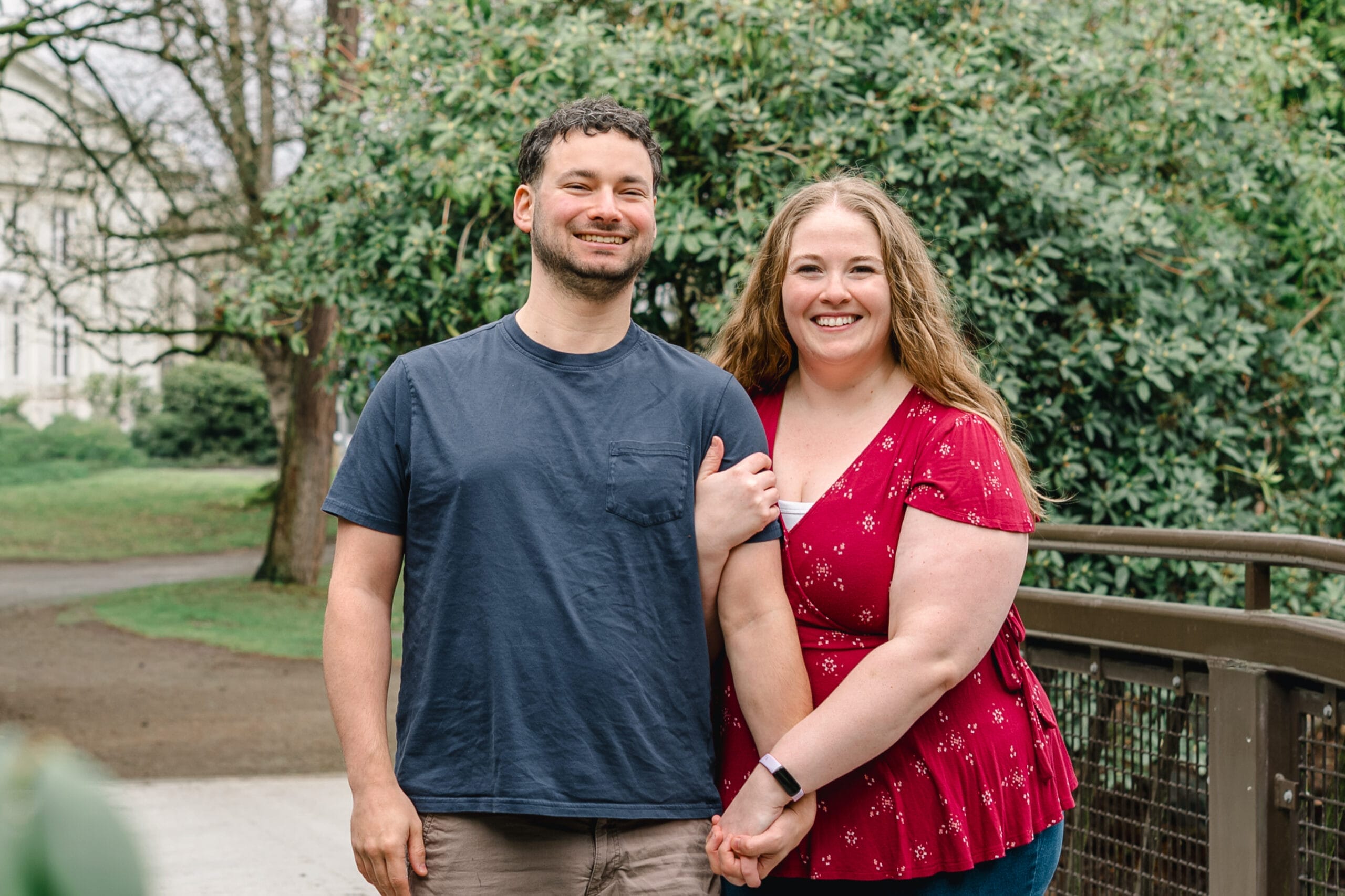 Couple standing side by side outdoors, smiling, with green trees and a wooden railing in the background.