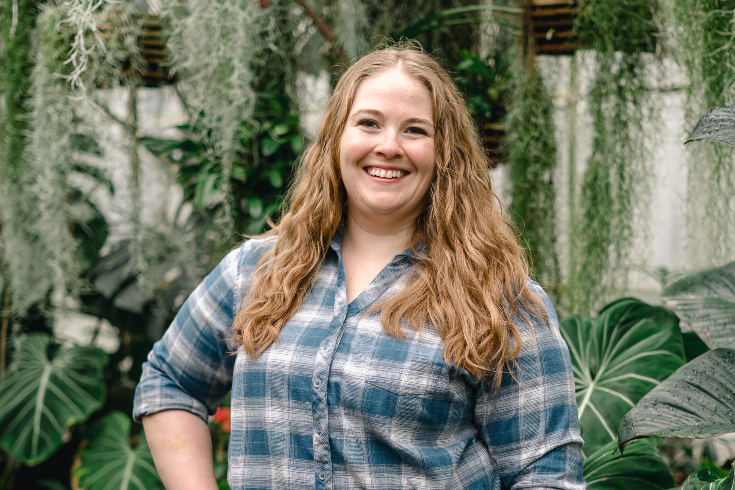 Smiling woman with long wavy hair in a blue plaid shirt, standing among lush green plants. (informative)