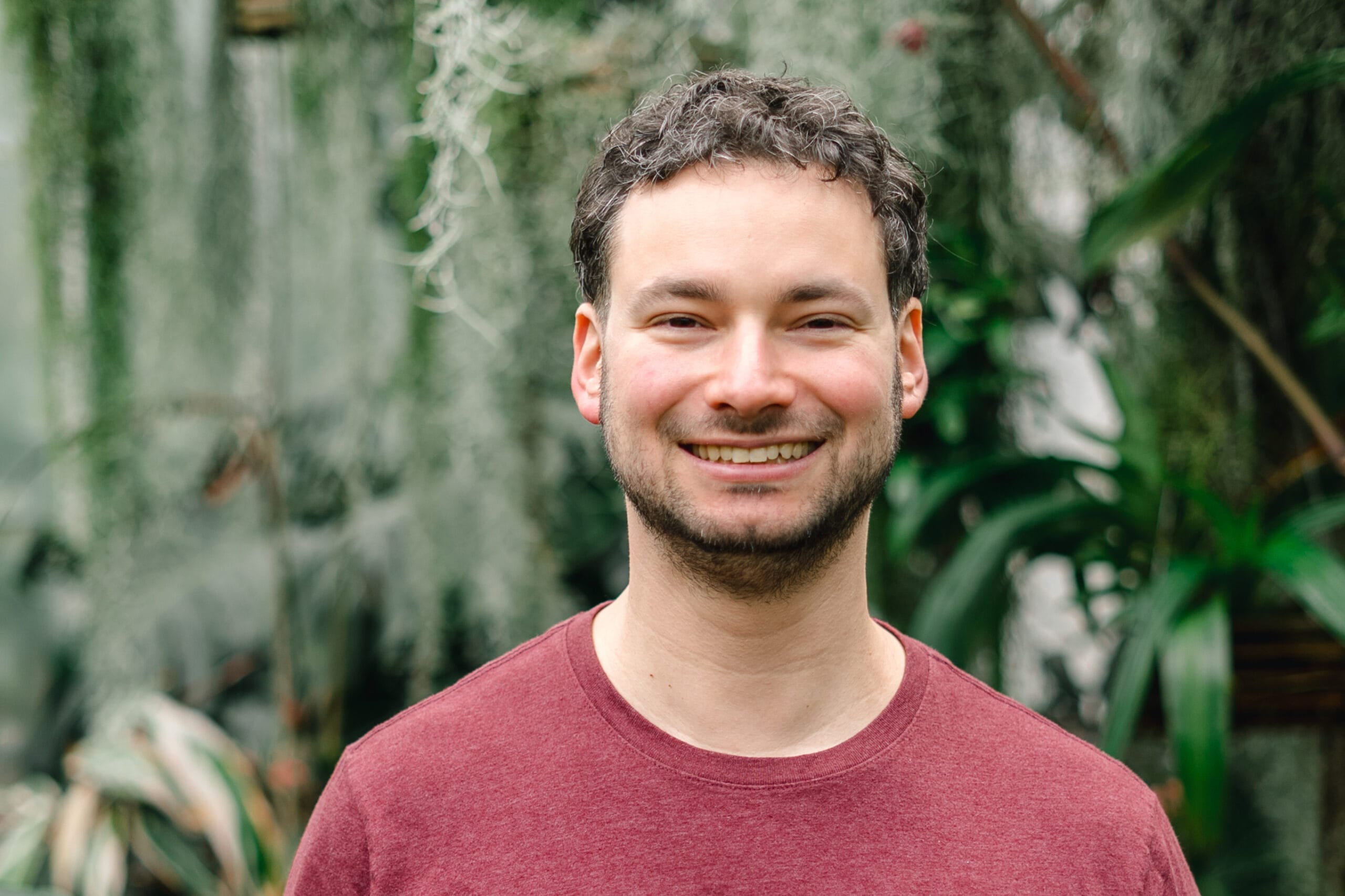 Smiling man in a maroon shirt outdoors with lush green plants behind him.