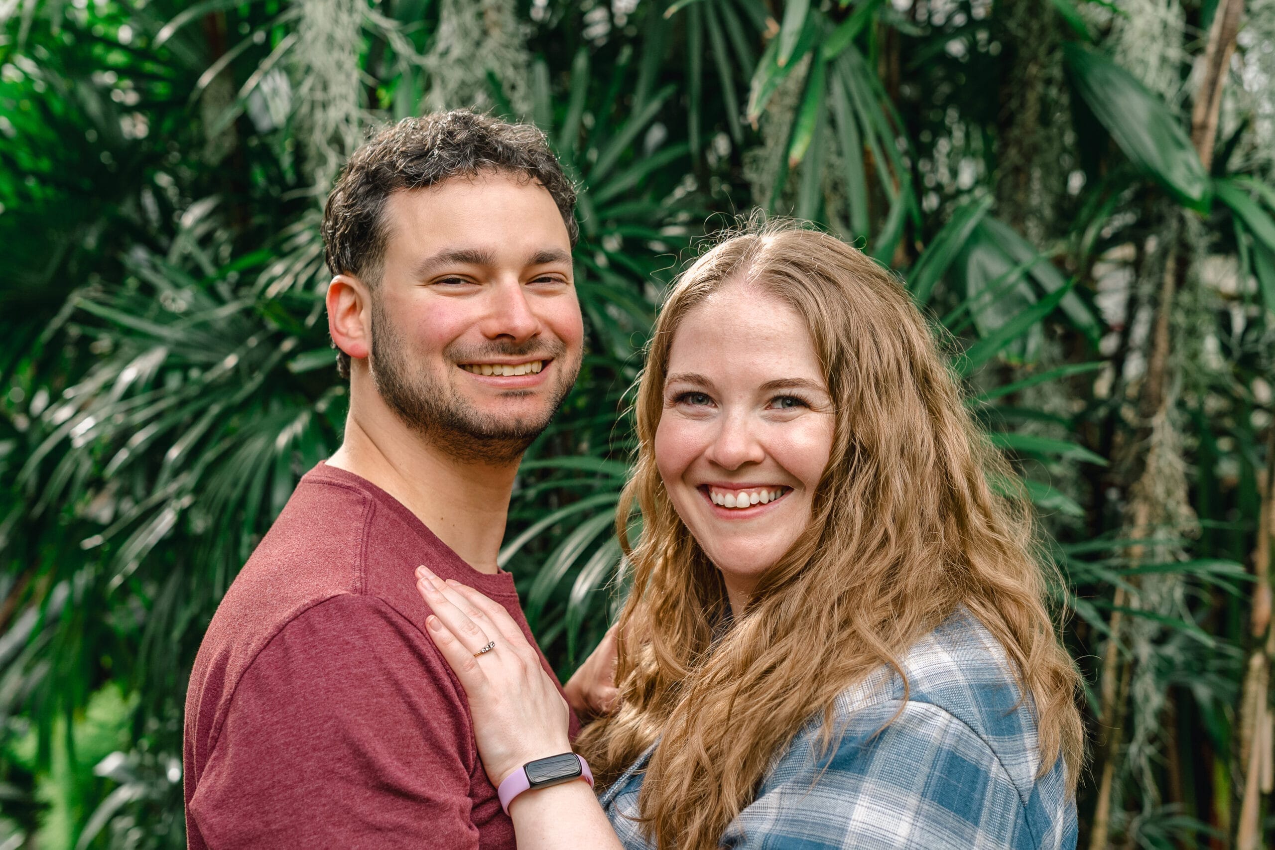 Smiling couple posing together outdoors in a lush green garden setting, arms around each other.