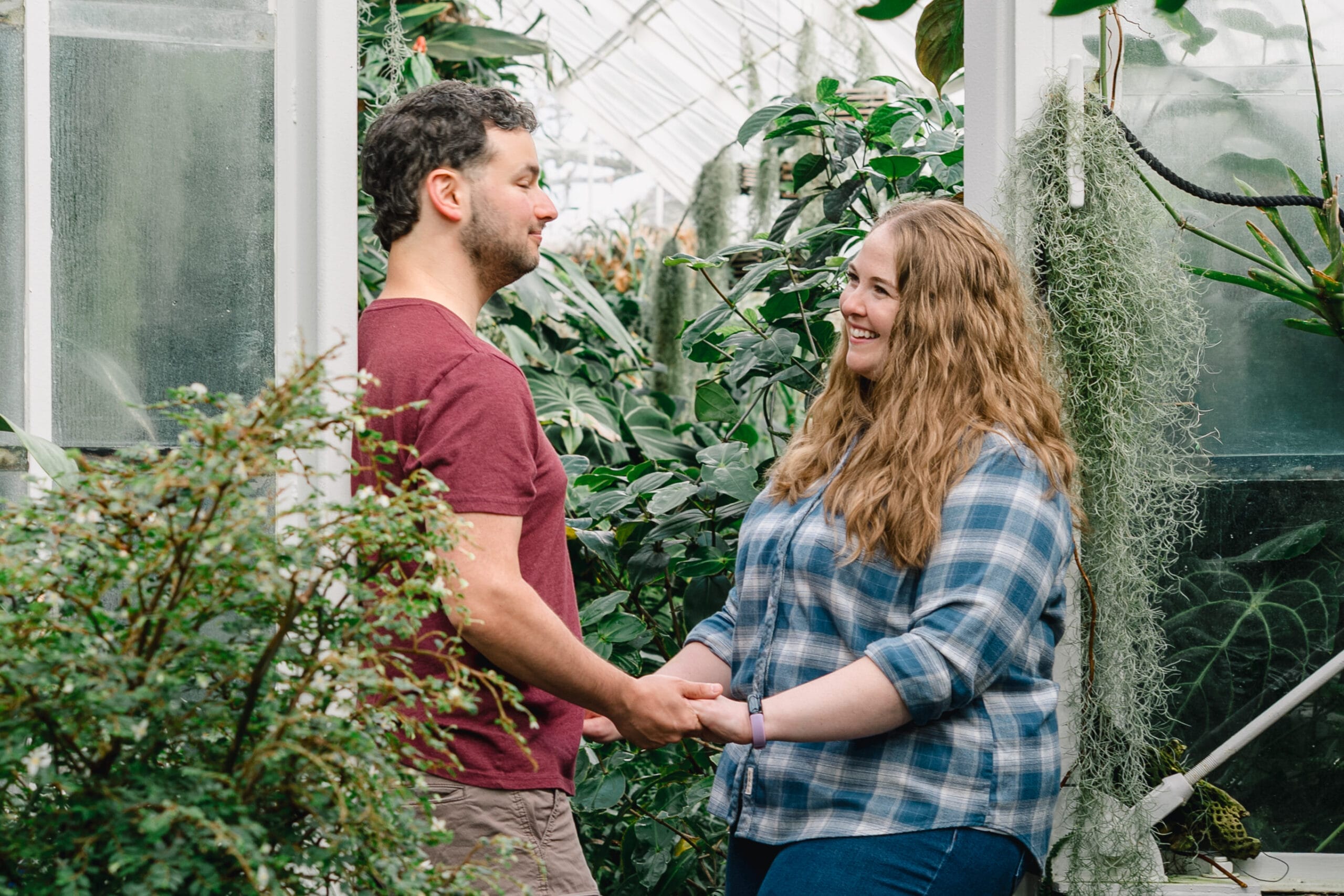 Couple standing in a greenhouse, holding hands and smiling at each other among lush plants.