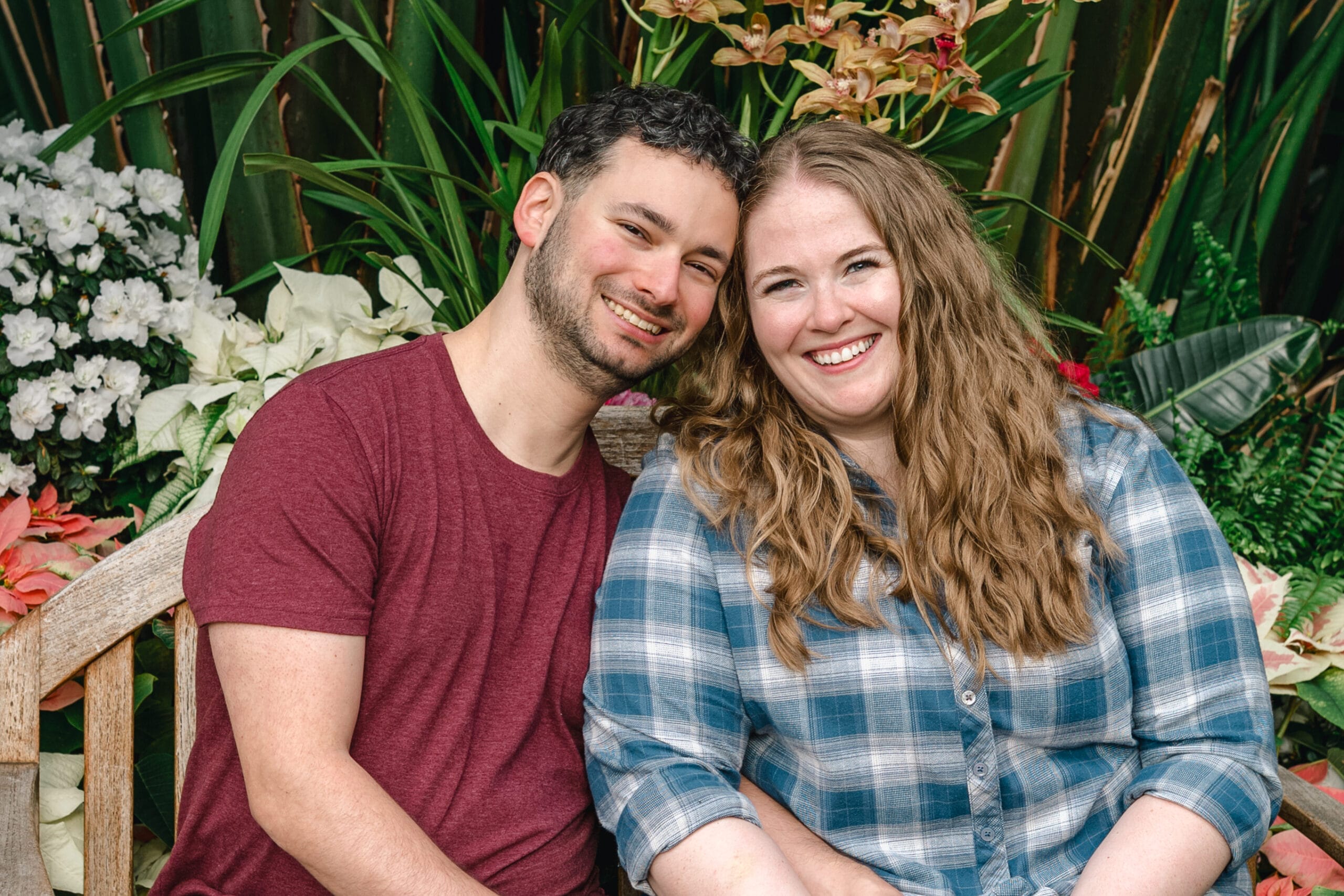 Smiling couple sitting close together on a wooden bench in a lush garden with white flowers and large green leaves behind them.