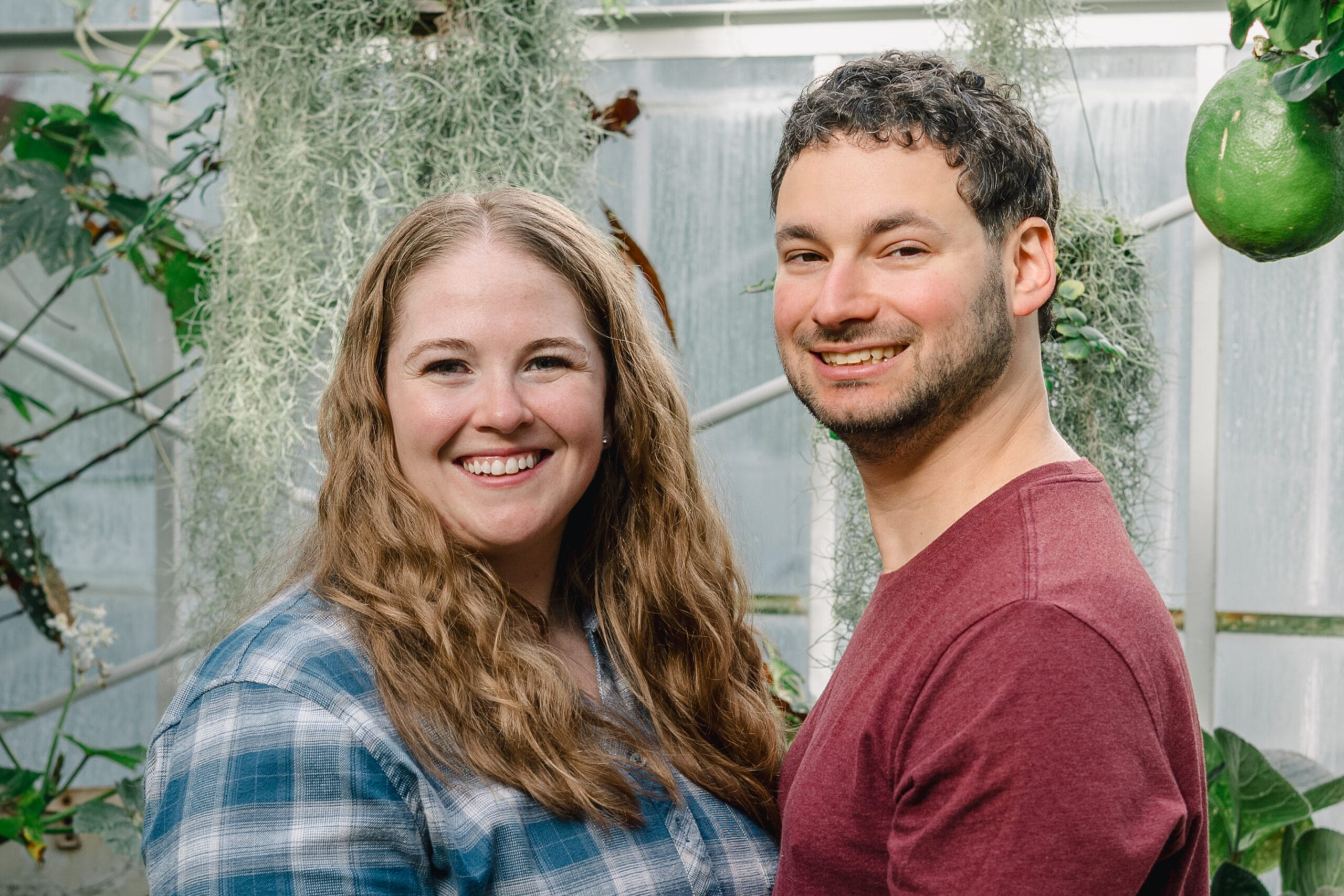 Two people smiling and posing together in a greenhouse with lush plants in the background.