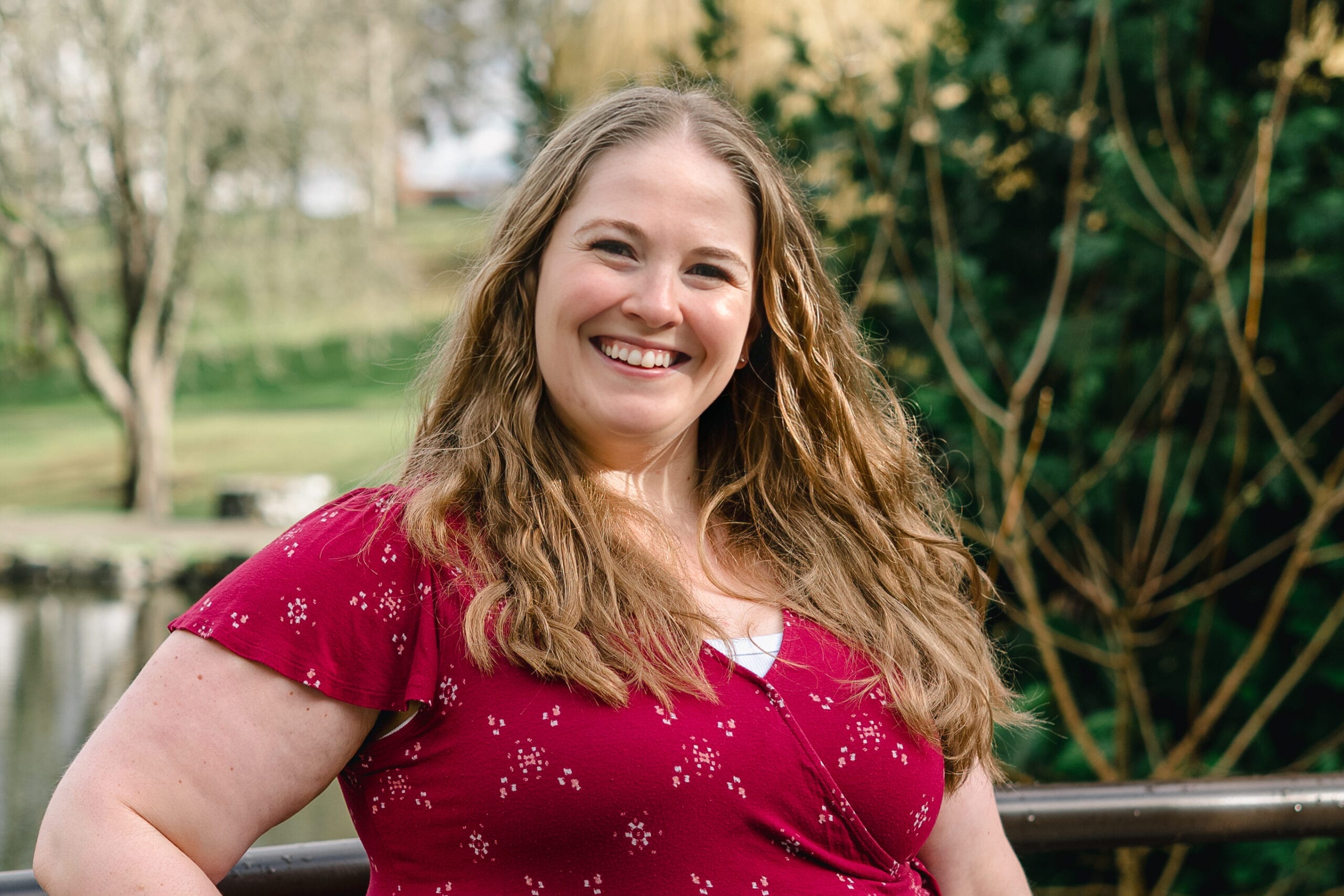 Smiling woman with long wavy hair wearing a red speckled top, outdoors near a railing and greenery behind her.