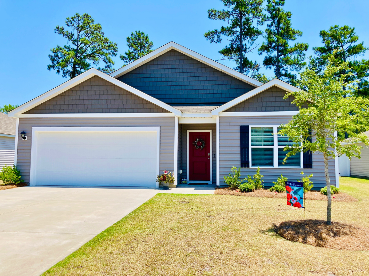 Front view of a single-story gray house with a two-car garage and red front door with wreath, a small yard and a welcome sign in the flower bed.
