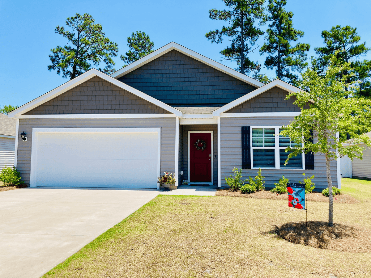 Front view of a gray, two-car garage house with a red front door and wreath, blue shutters, and a small welcome sign in the yard.