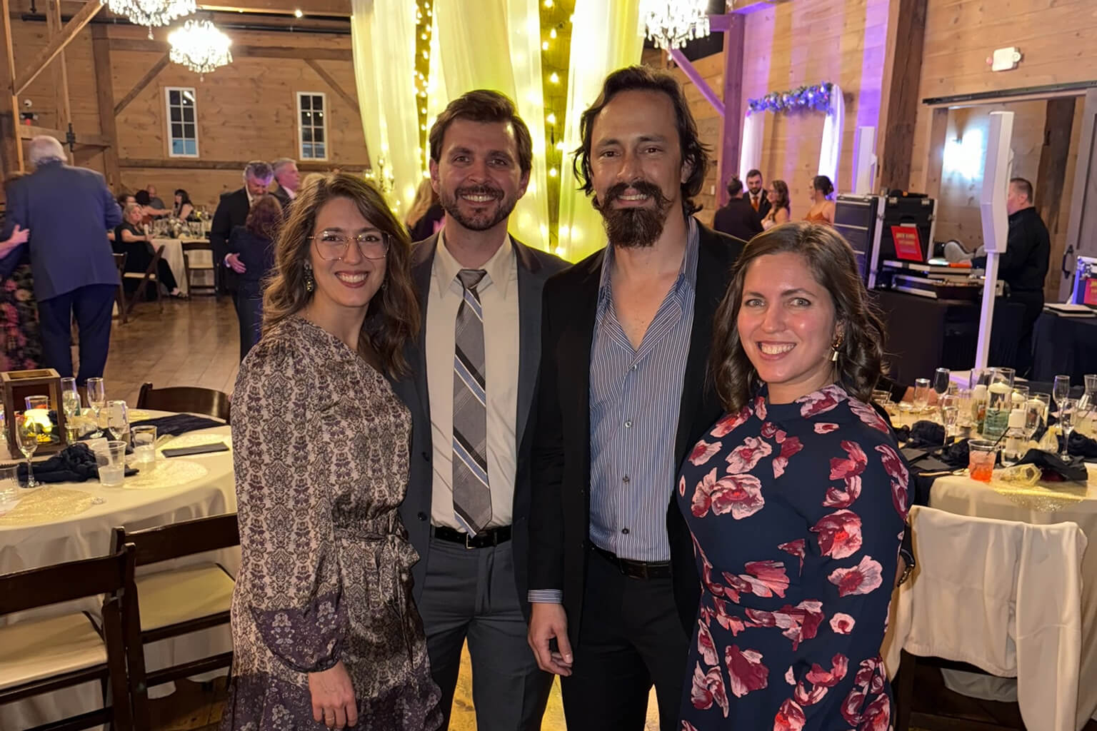 Group of four adults smiling for a photo at a decorated indoor wedding reception with chandeliers and string lights in the background.