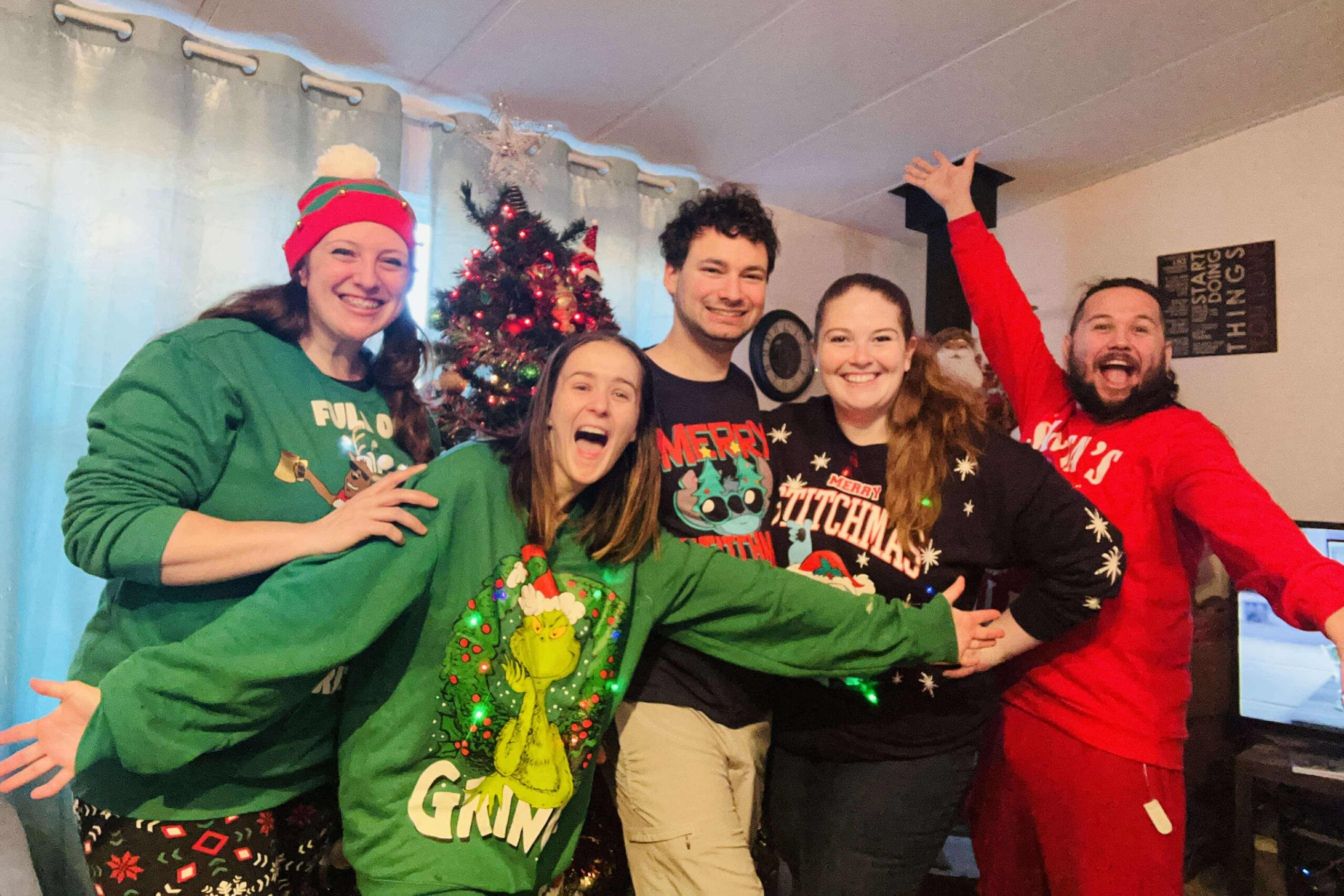 Group of people in festive Christmas sweaters smiling with arms outstretched indoors, a Christmas tree in the background.