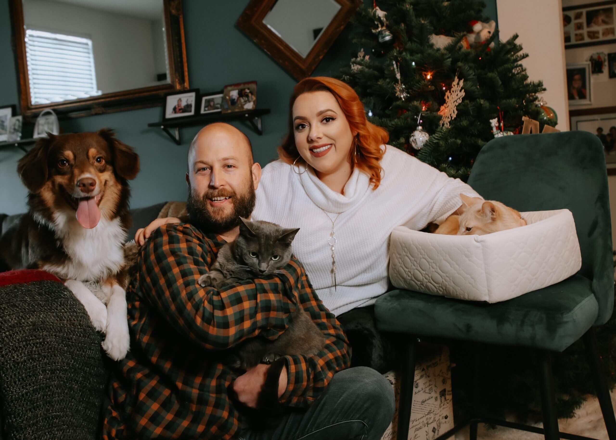 Smiling couple on a couch with two dogs and a cat, Christmas tree in the background.