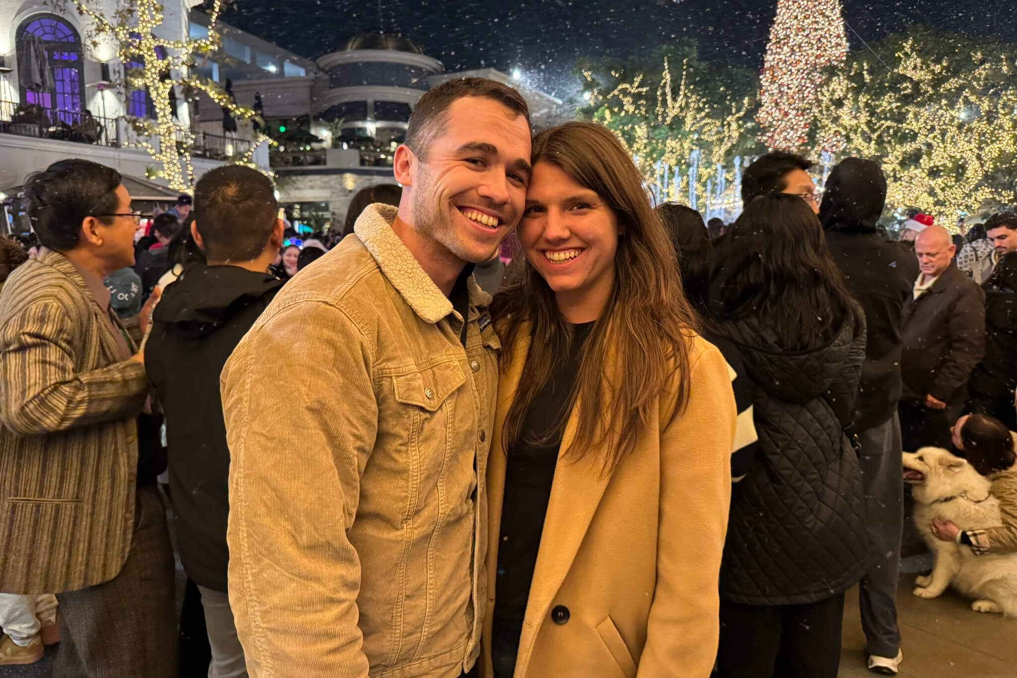 Smiling couple posing at a Christmas light festival with twinkling trees and a crowd behind them.