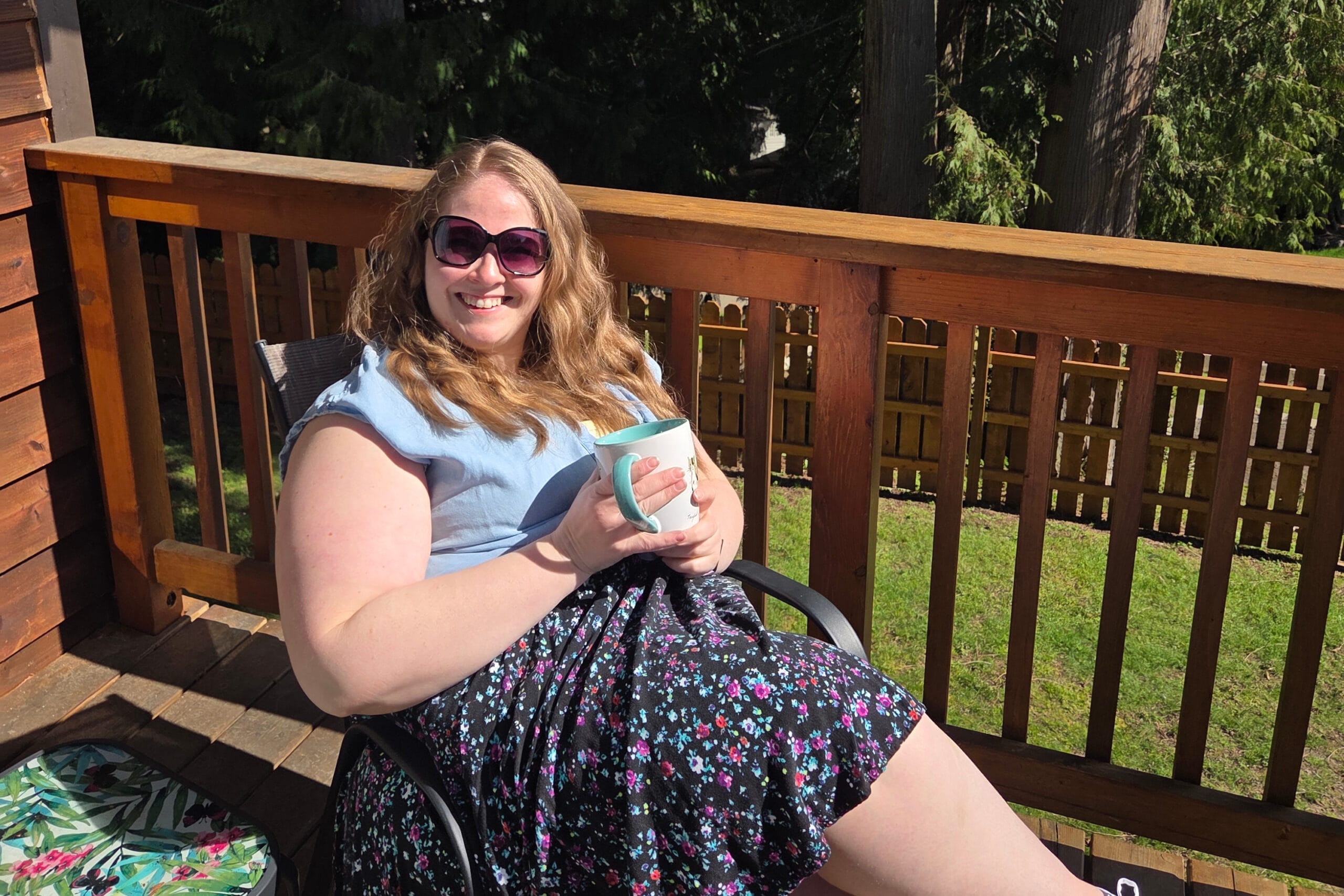 Woman smiling on a wooden deck outdoors, wearing sunglasses and a blue top, holding a teal mug with a floral skirt in sunlight.