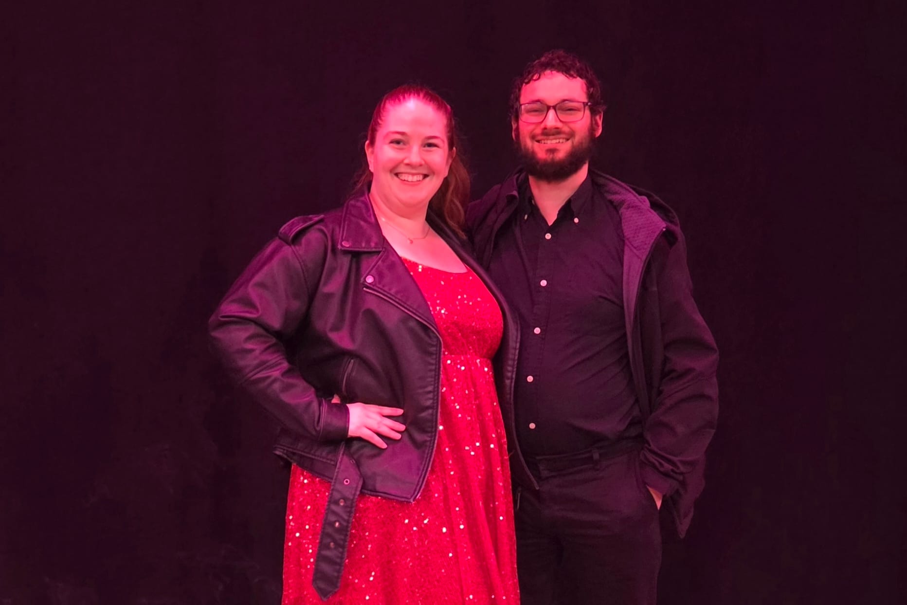 Two people posing together in pink stage lighting, smiling; woman in a red sequin dress and black leather jacket, man in a black shirt and jacket.