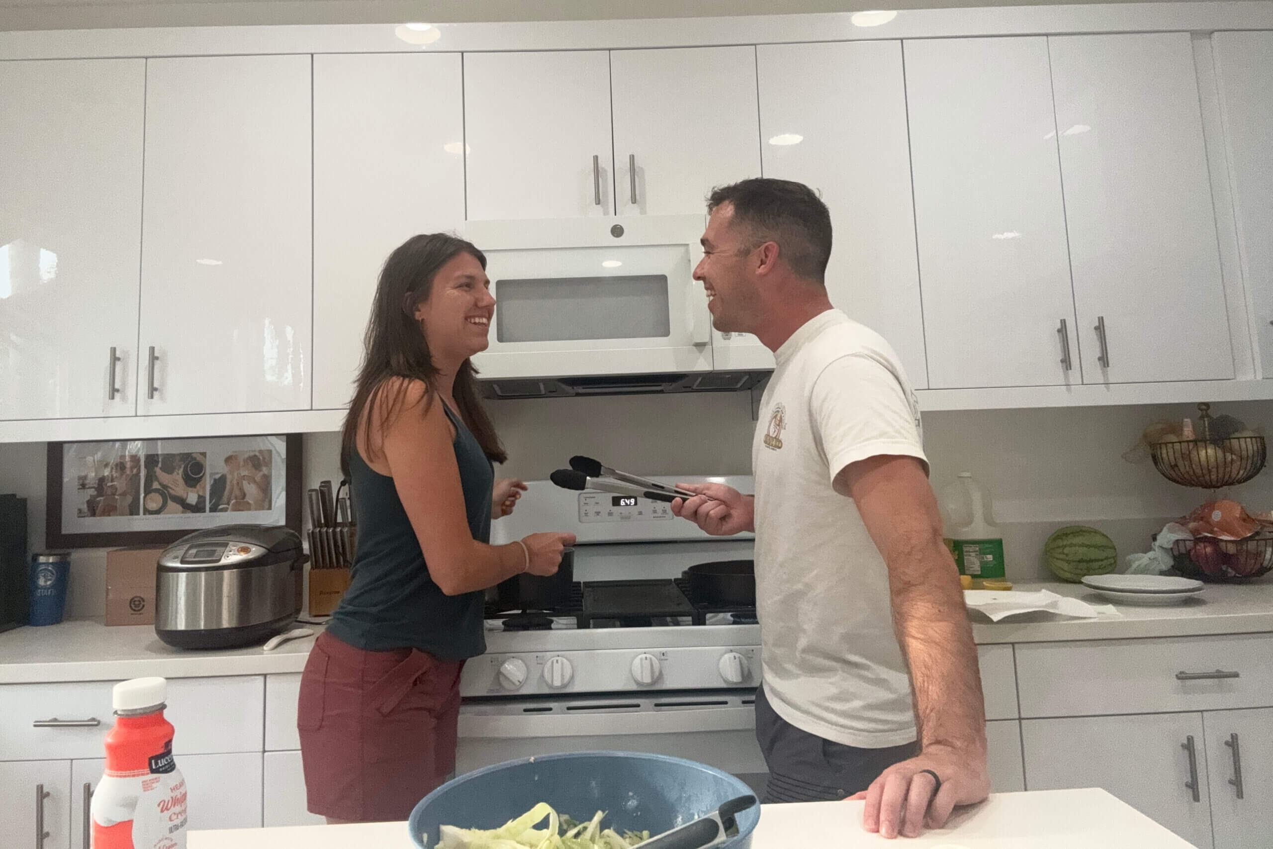 Two adults smiling and facing each other over a kitchen counter by a gas stove, as they prepare food together.