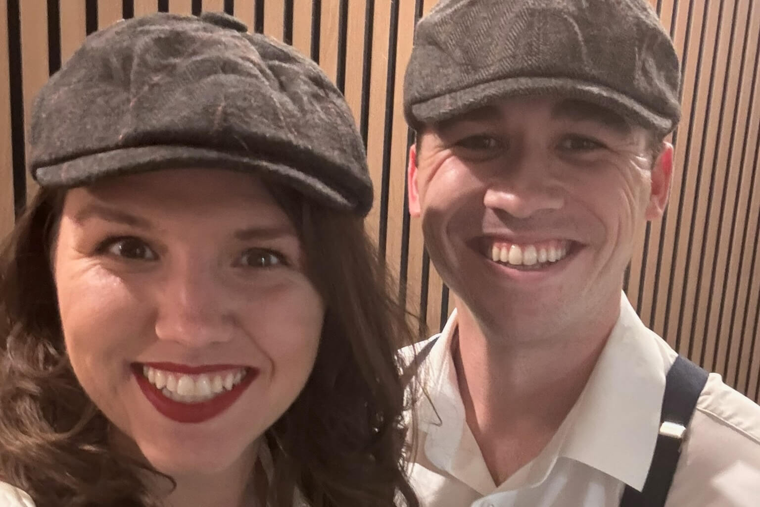 Close-up of a woman and a man wearing gray flat caps, smiling at the camera in front of a wood-paneled wall; she has red lipstick and wavy hair, he wears a light shirt with suspenders.