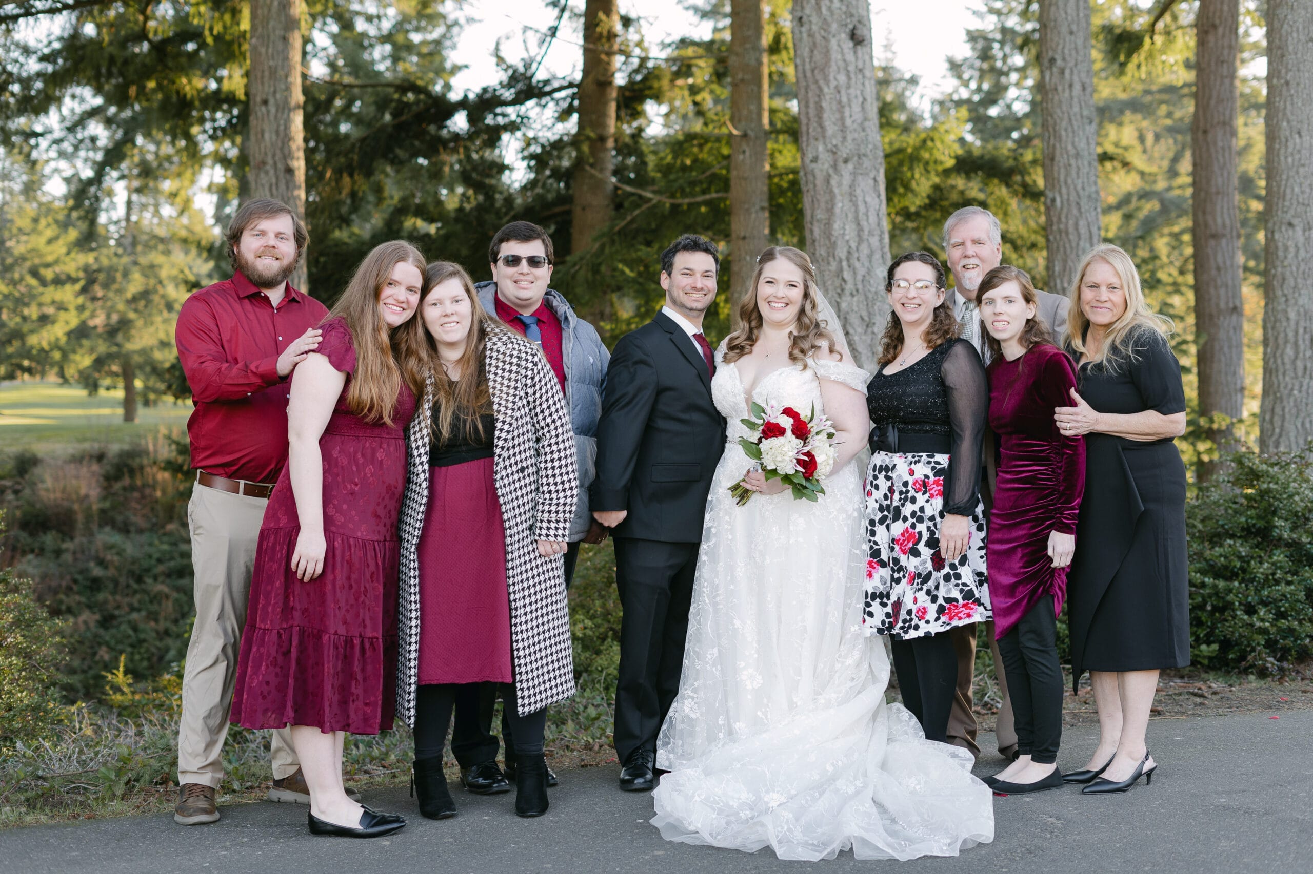 Wedding party photo: bride and groom center, surrounded by family and friends outdoors among trees, everyone smiling.