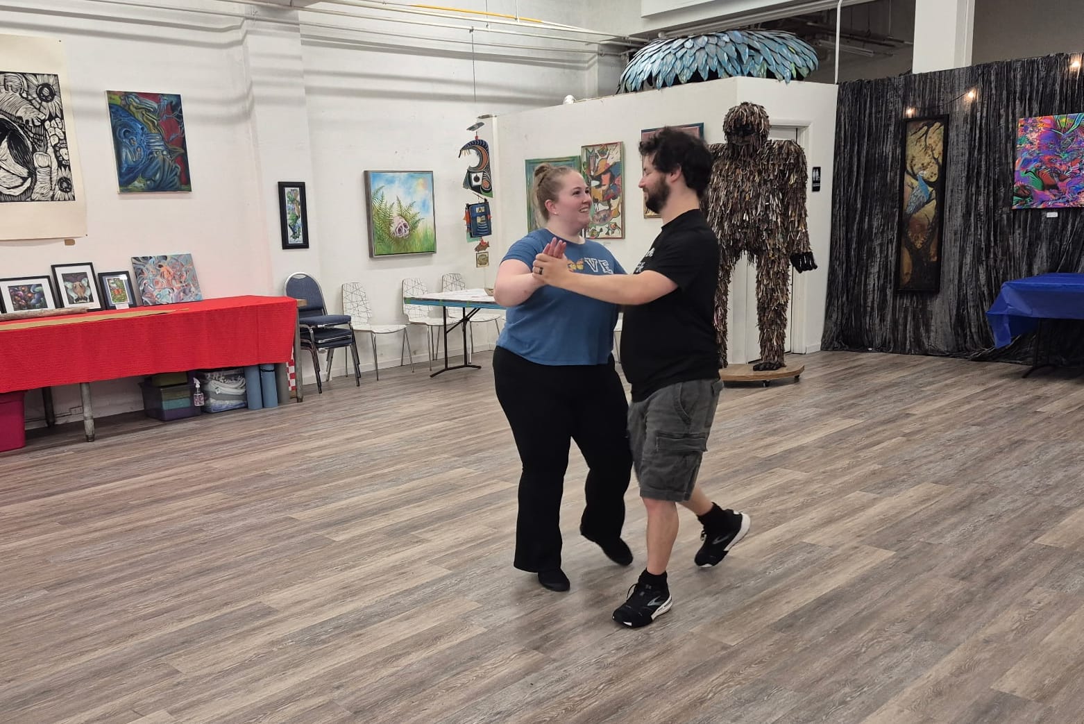 Couple dancing together in an art gallery with paintings on the walls and a large feather sculpture overhead.