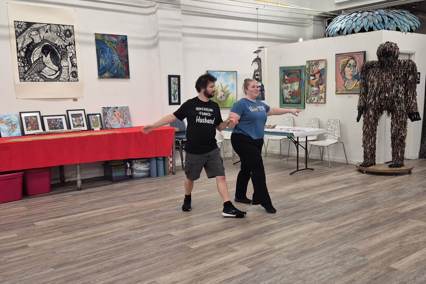 Man and woman dancing in an art studio, holding hands as they sway; colorful paintings line the walls and a large shaggy sculpture stands to the right.