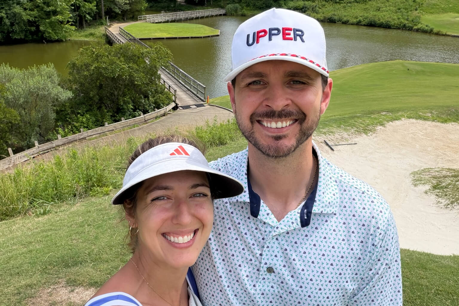 Smiling couple taking a selfie on a grassy golf course beside a pond and wooden bridge.