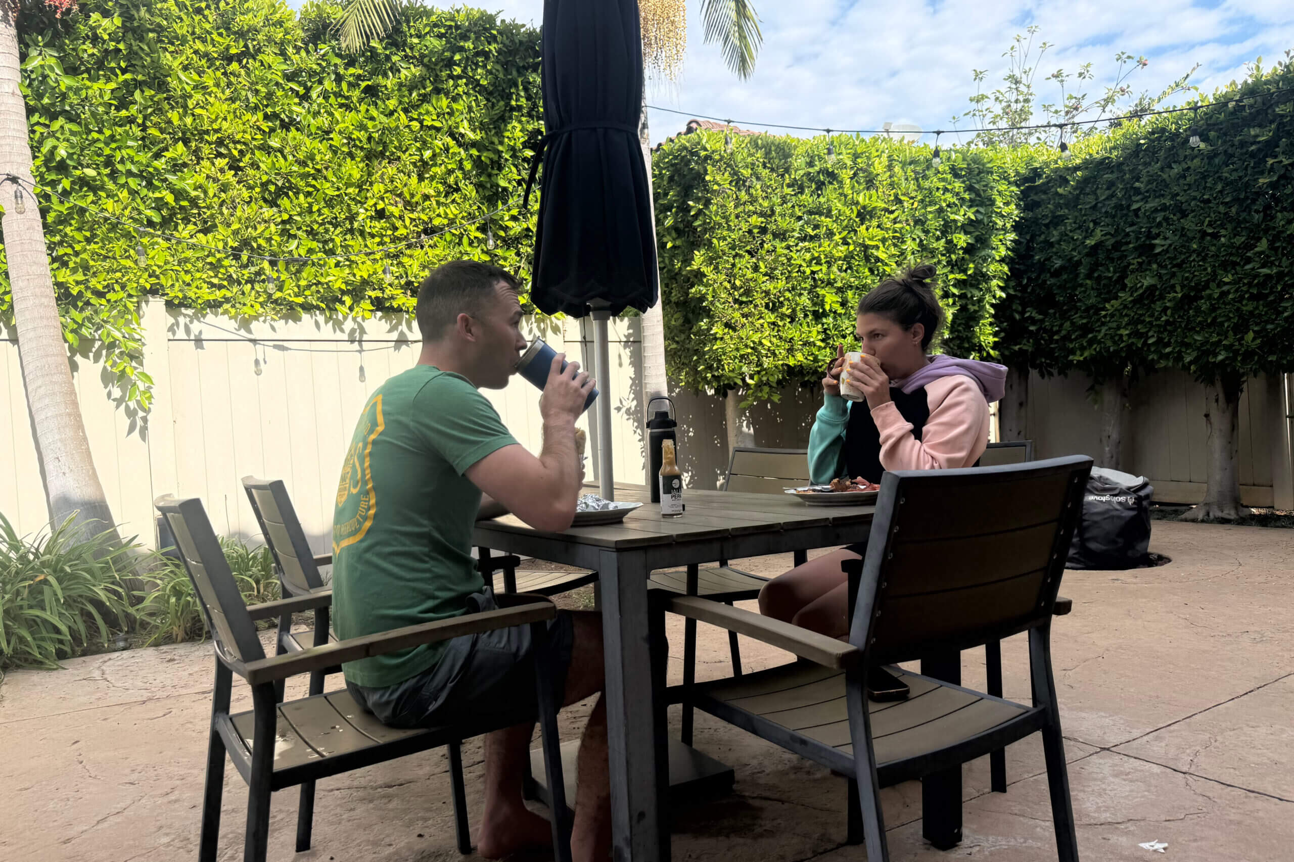 Man in green shirt and a woman in a pink hoodie sit at an outdoor patio table drinking from mugs, with string lights and green hedges in the background.