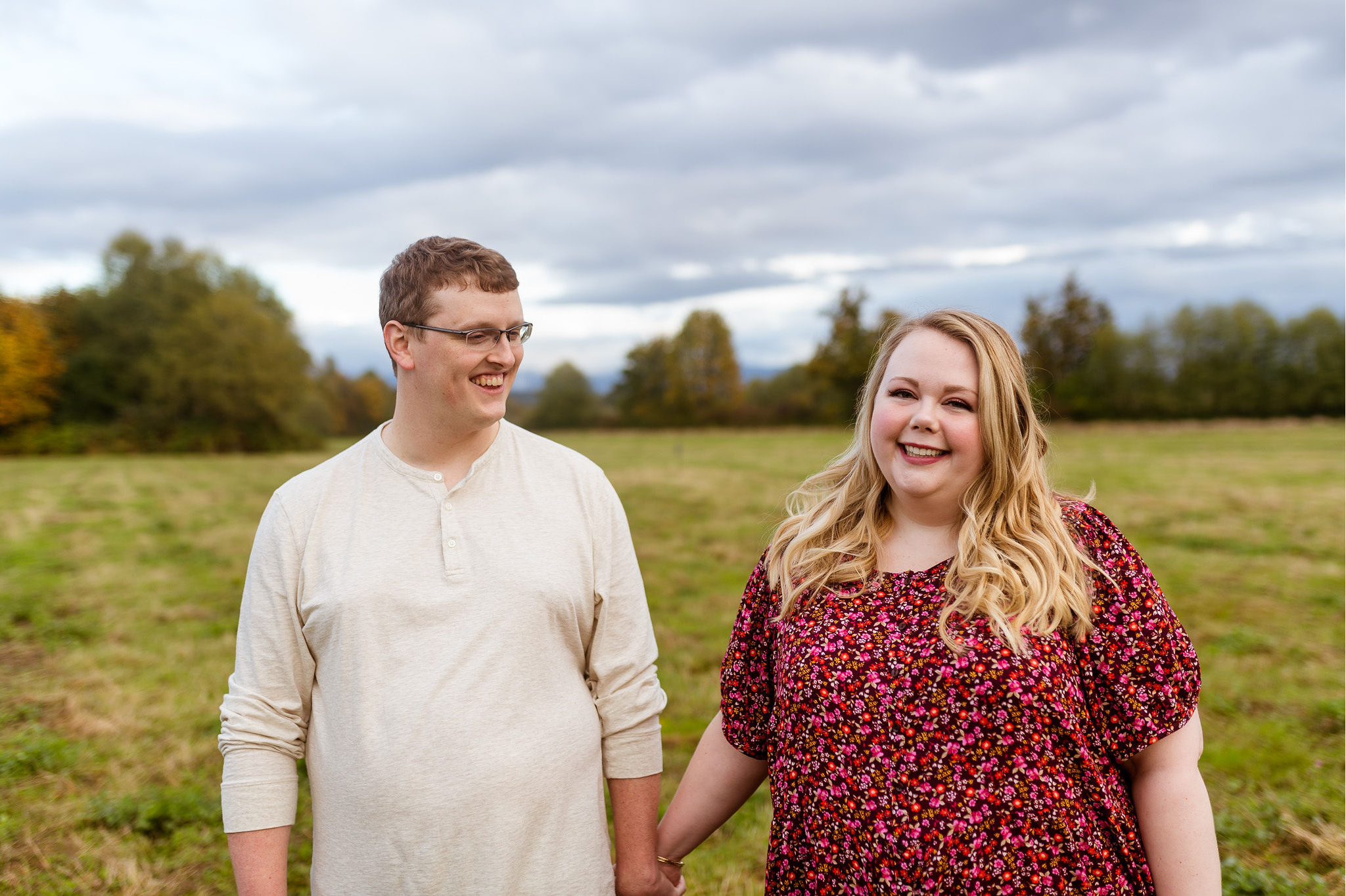 Smiling couple standing in a green field with trees and a cloudy sky above them.