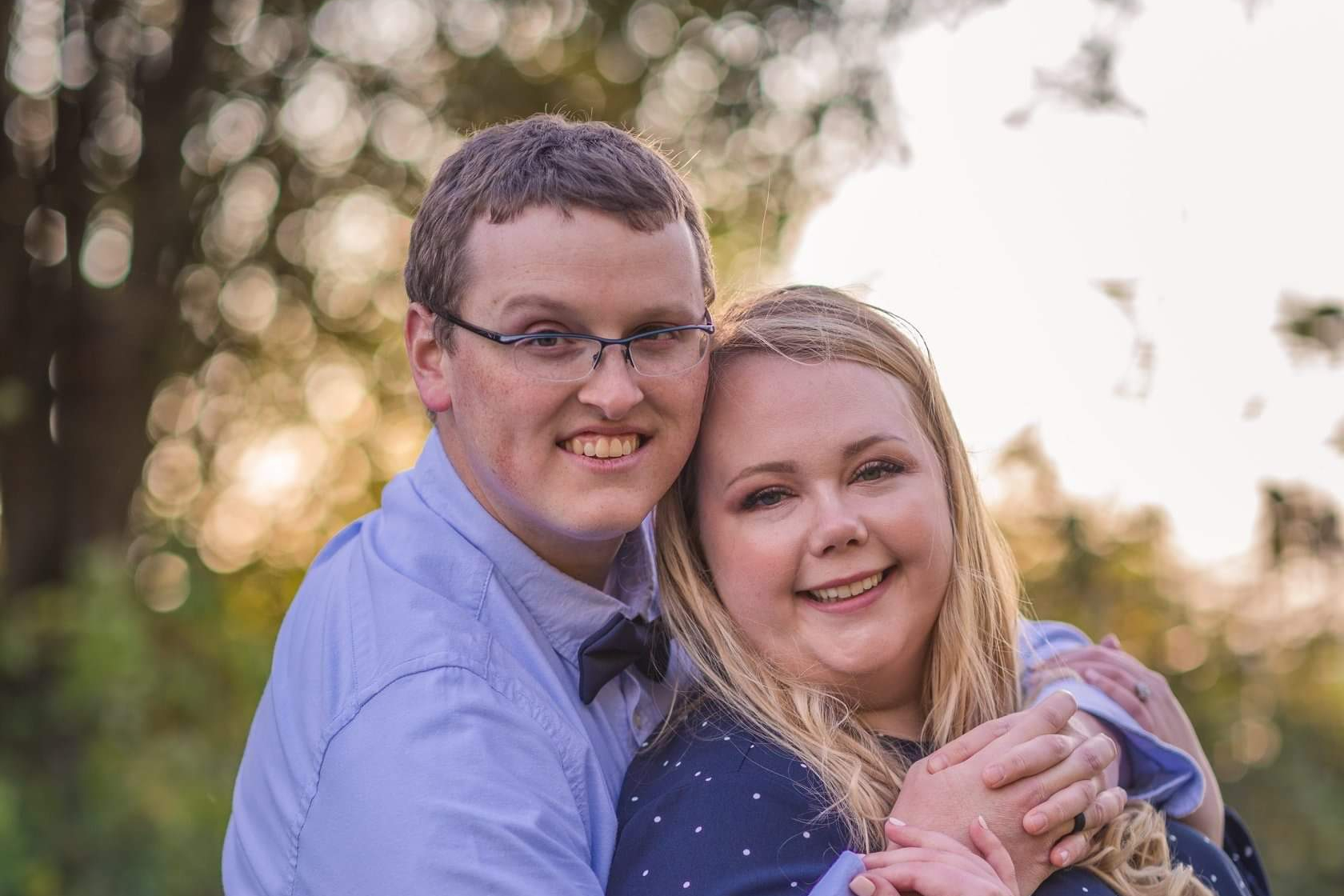 Couple embracing and smiling at the camera in a sunny park setting with blurred trees in the background.