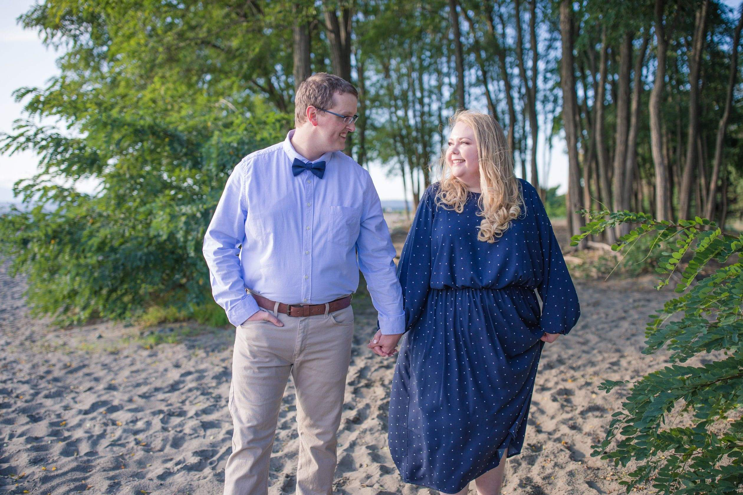 Couple walking hand in hand along a sandy beach, smiling at each other with trees in the background