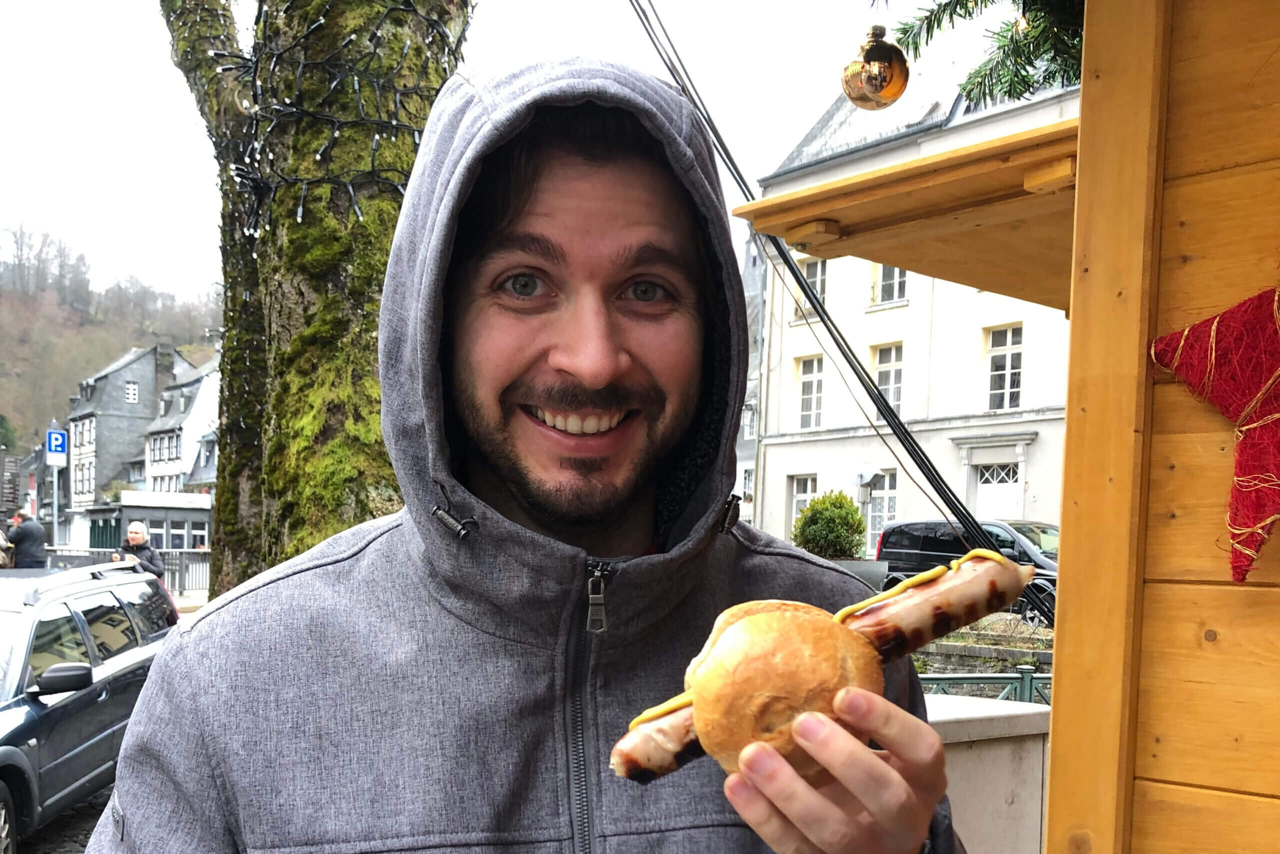 Man in a gray hooded jacket smiling while holding a hot dog at an outdoor market street stall nearby.