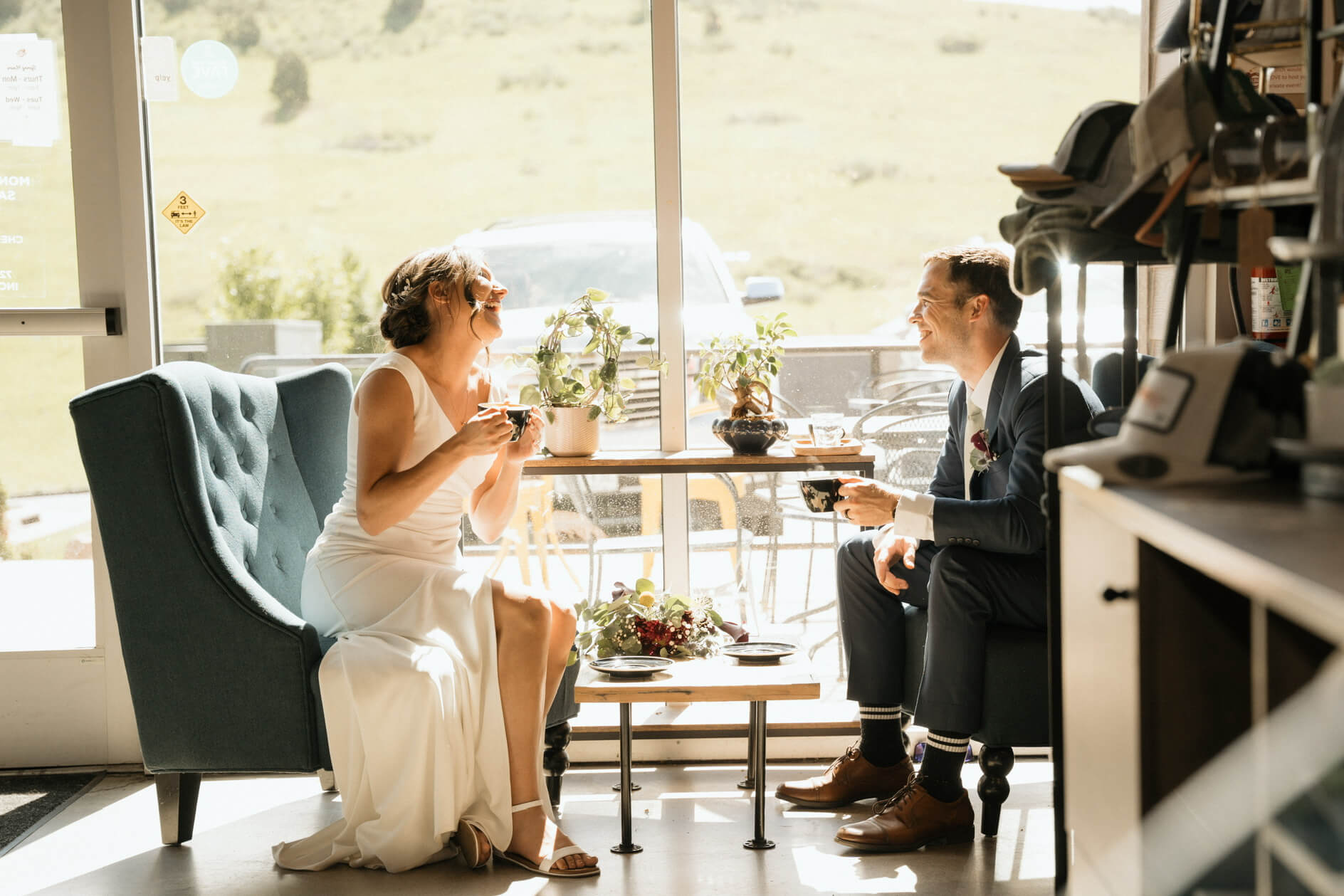 Couple in a sunlit cafe share a laugh over coffee; the bride in a white dress, groom in a suit sits across from her with plants on the table behind them.