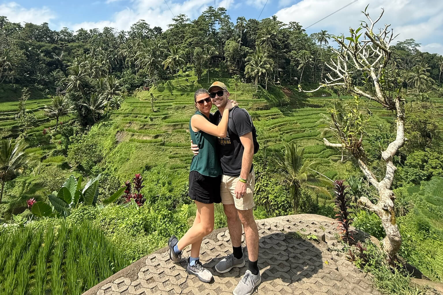A couple stands on a stone platform hugging, with lush terraced rice fields and palm trees behind them.