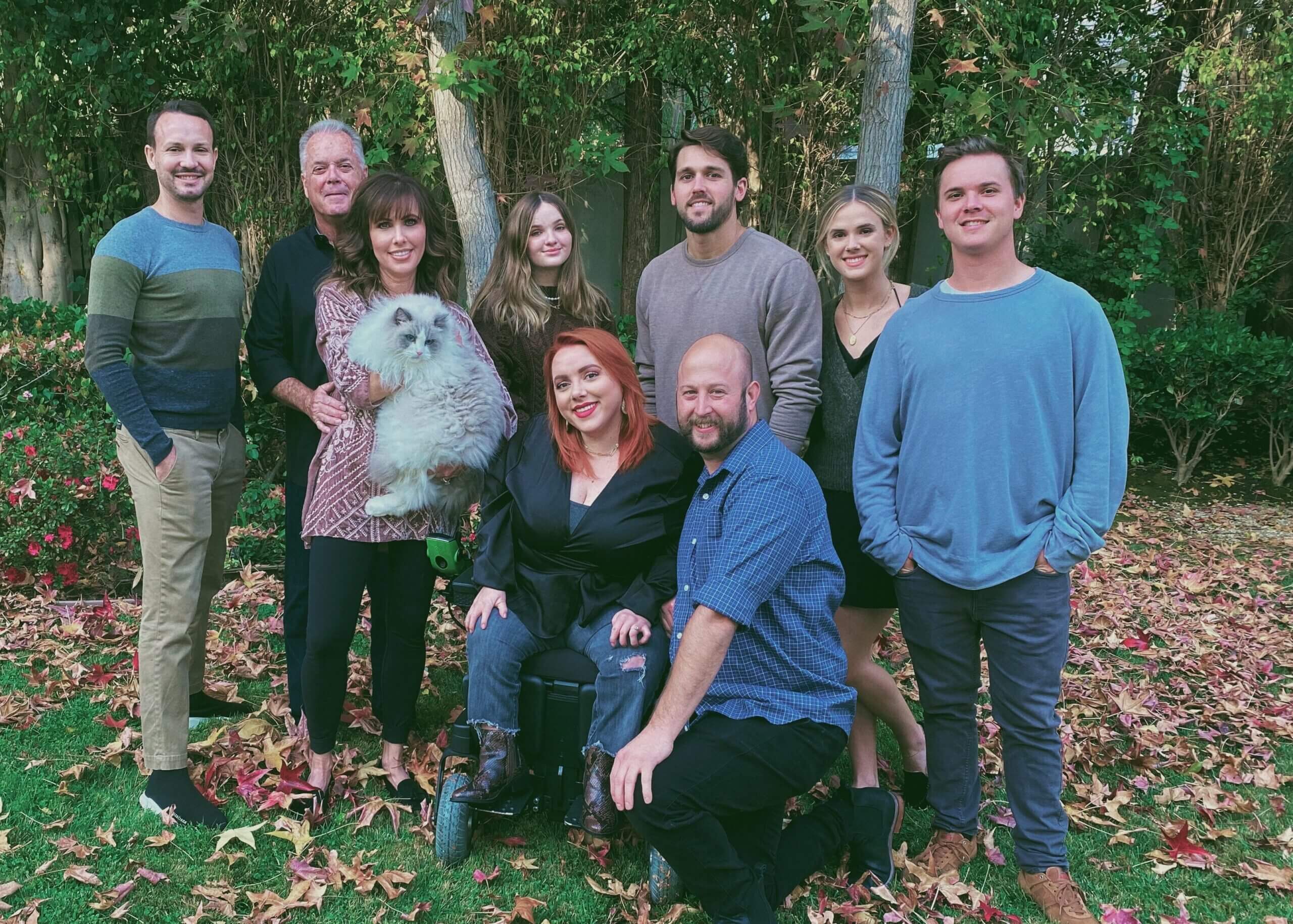 Group of adults posing outdoors in a yard with autumn leaves; a woman holds a fluffy white dog in front of a shrubbery.
