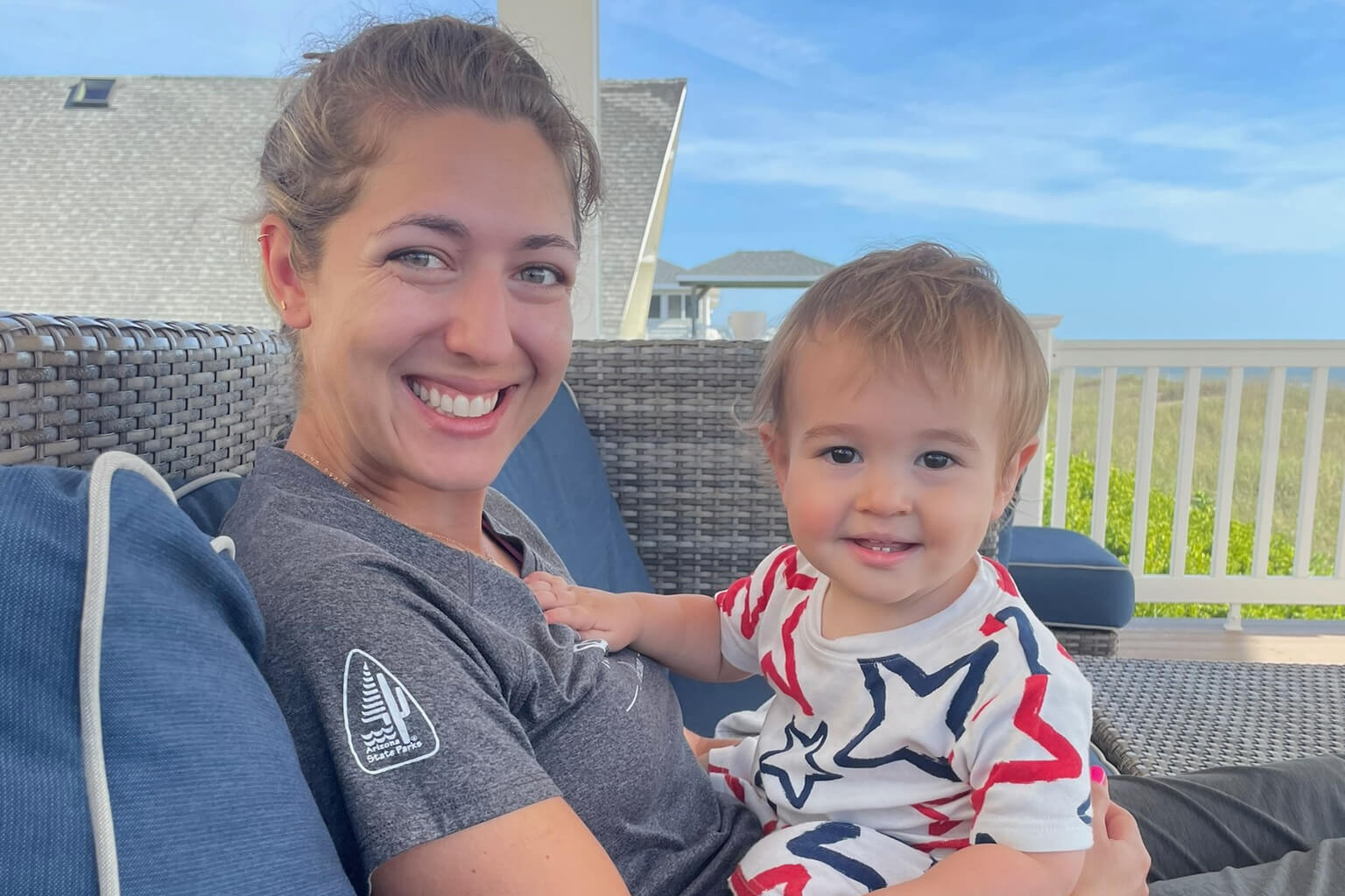 Smiling woman and toddler on a wicker outdoor couch with a blue sky and white railing behind them.