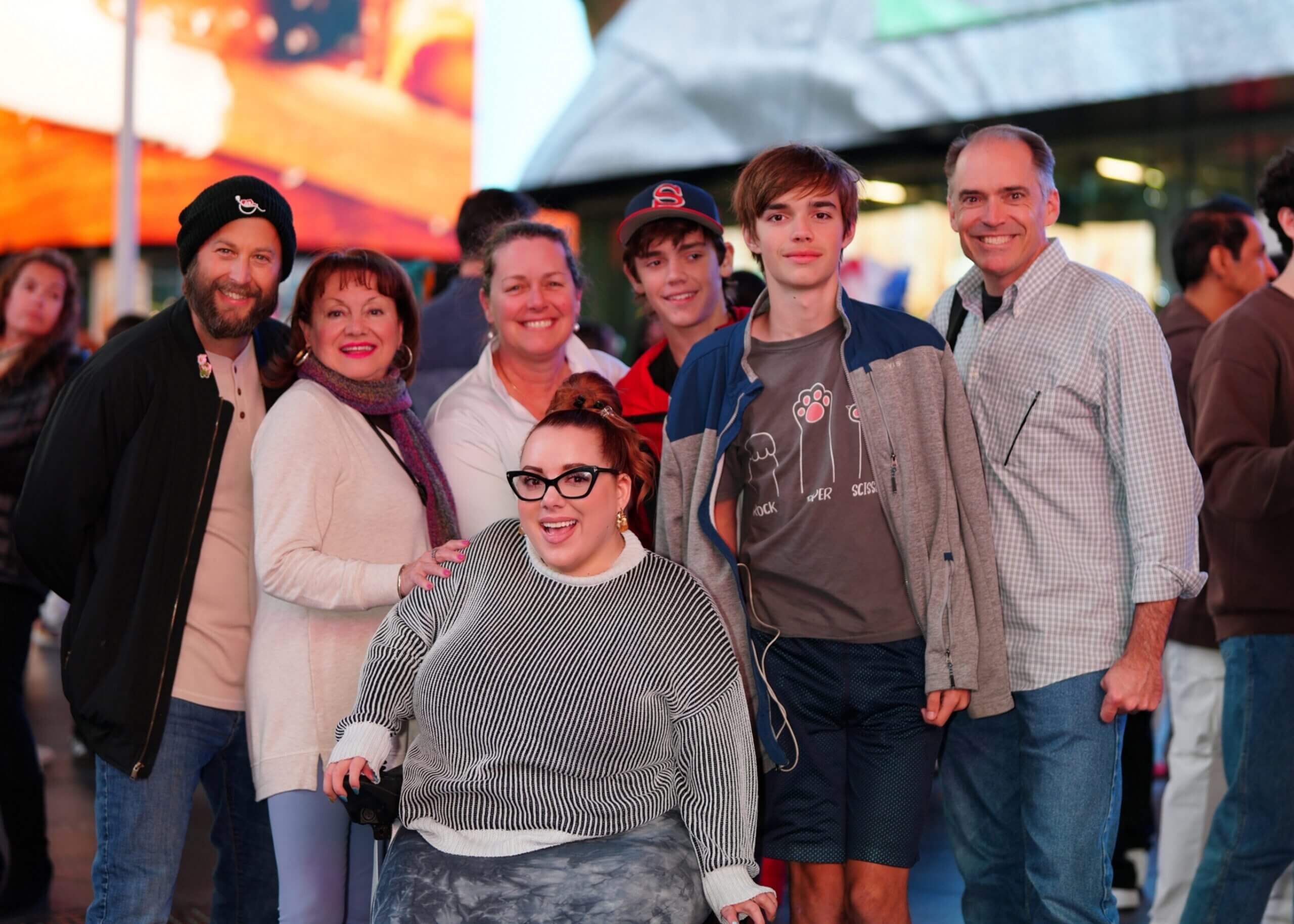 Eight smiling people posing together outdoors in an urban setting with colorful lights in the background, looking at the camera.