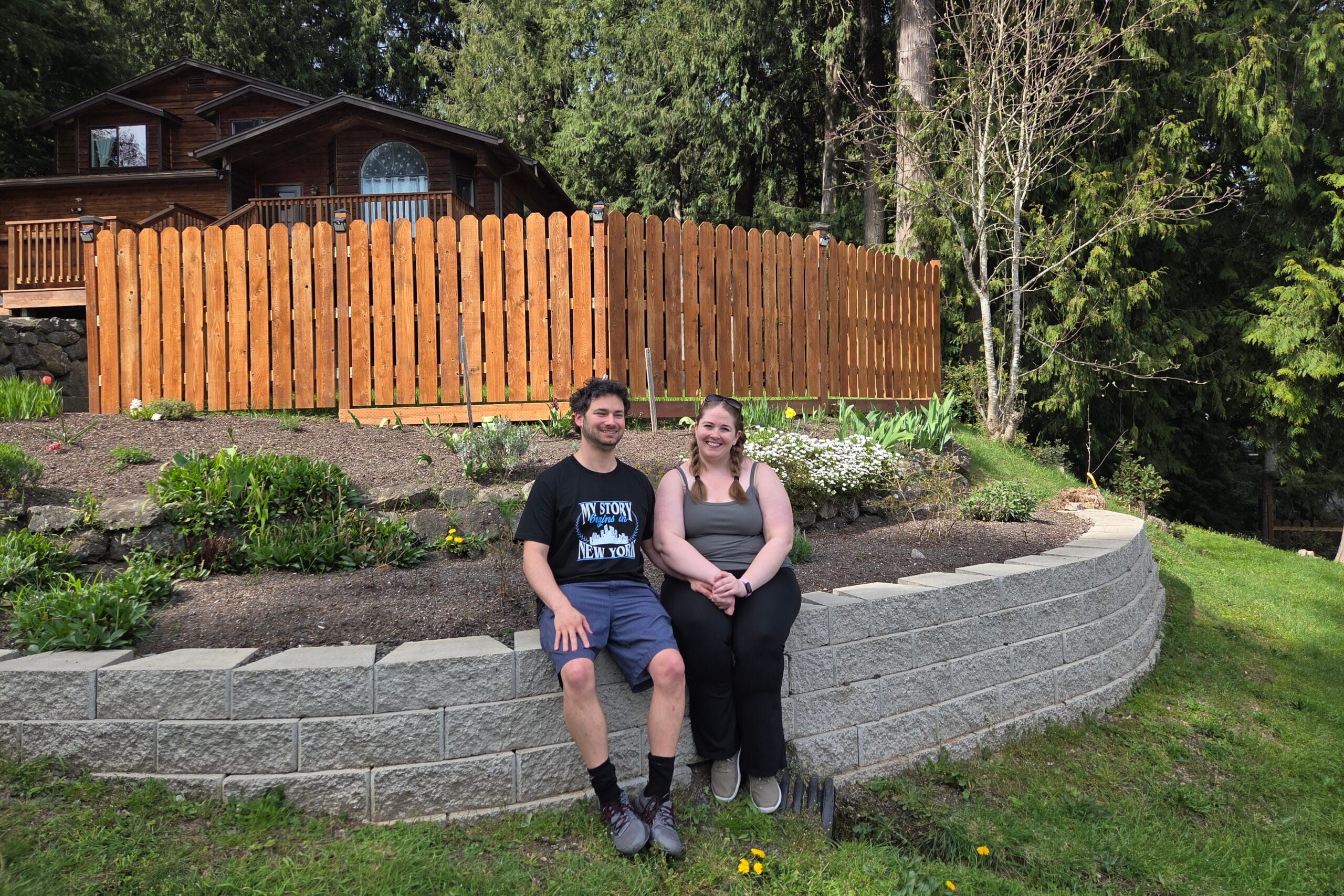 Smiling couple sitting on a stone retaining wall in a garden, with a tall wooden fence and forest behind them.