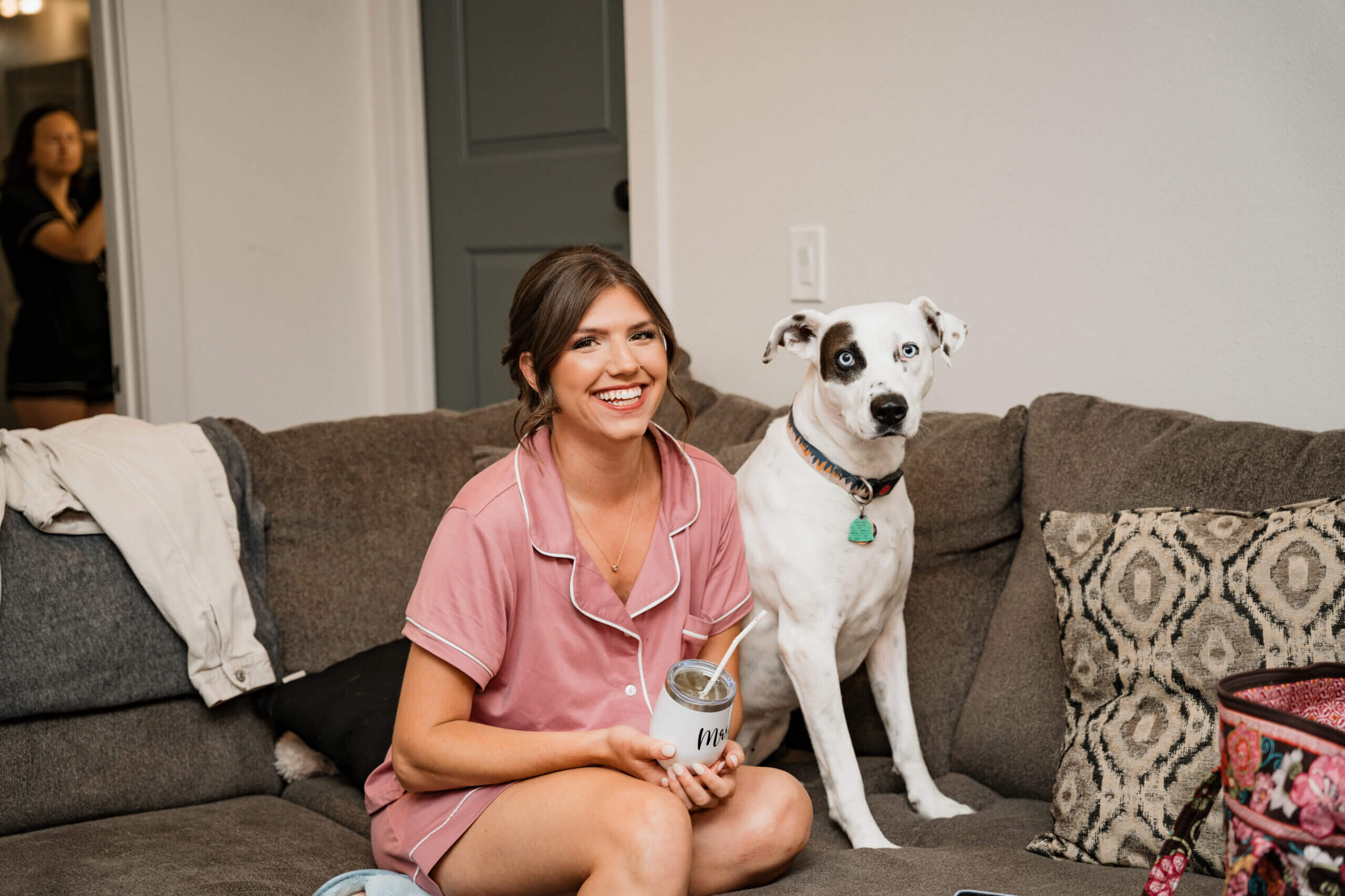 Smiling woman in pink pajamas sits on a gray couch beside a white dog with a black patch over one eye and blue eyes.