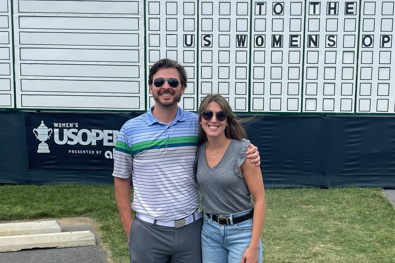Smiling couple wearing sunglasses stands arm-in-arm in front of a large scoreboard at a Women's US Open event.