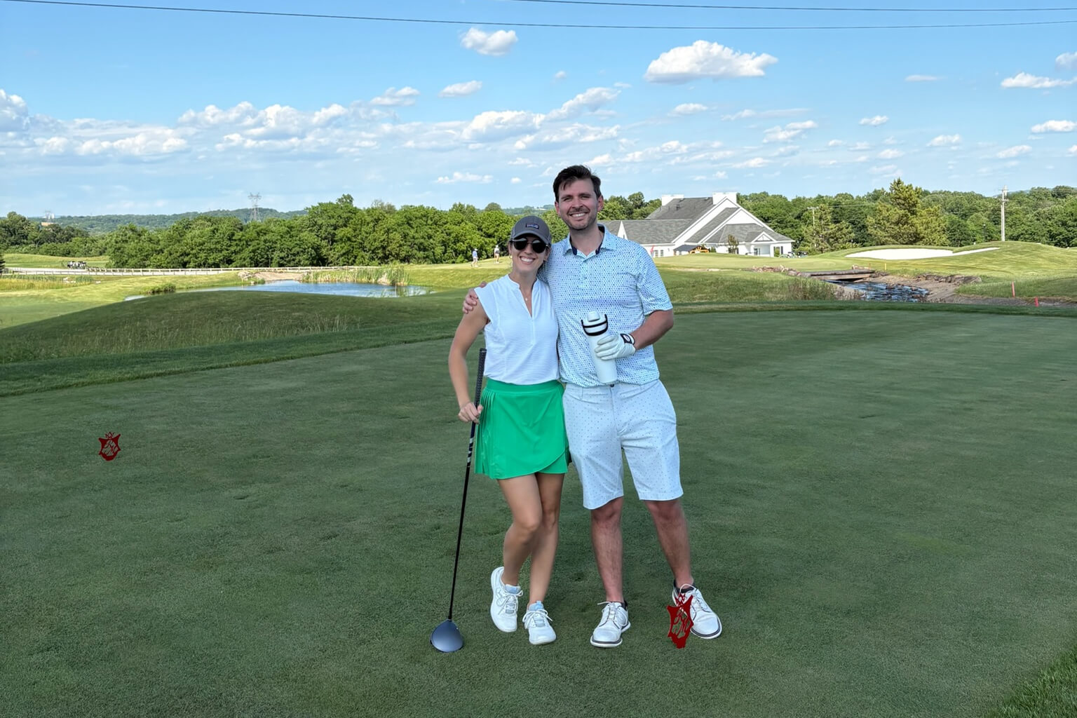 Smiling couple posing on a golf course green with clubs, a water feature, and a clubhouse in the distance on a sunny day.